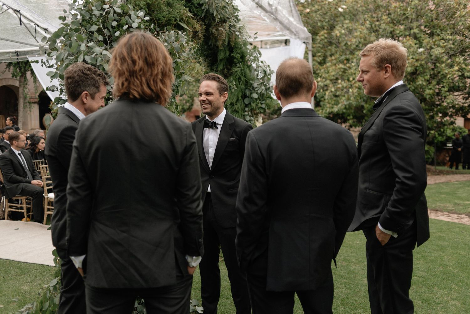 Groomsmen in black suits gather on a lawn during an outdoor wedding ceremony.