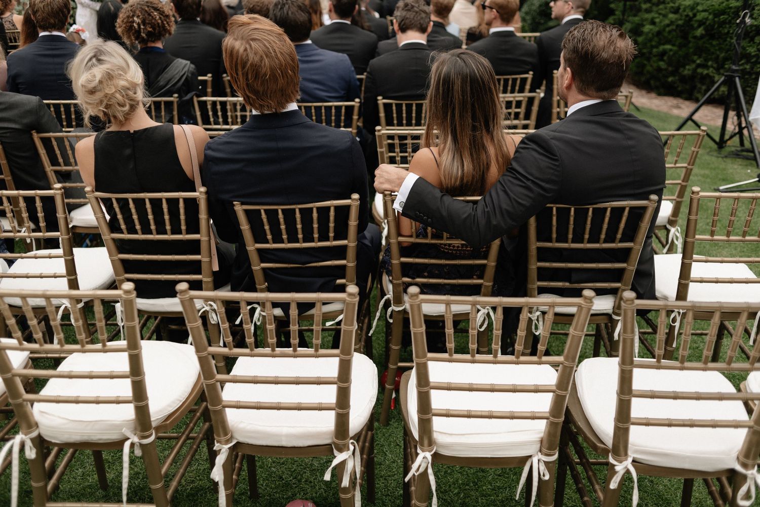 Empty white chairs arranged in rows behind seated guests at an outdoor wedding ceremony on green grass.