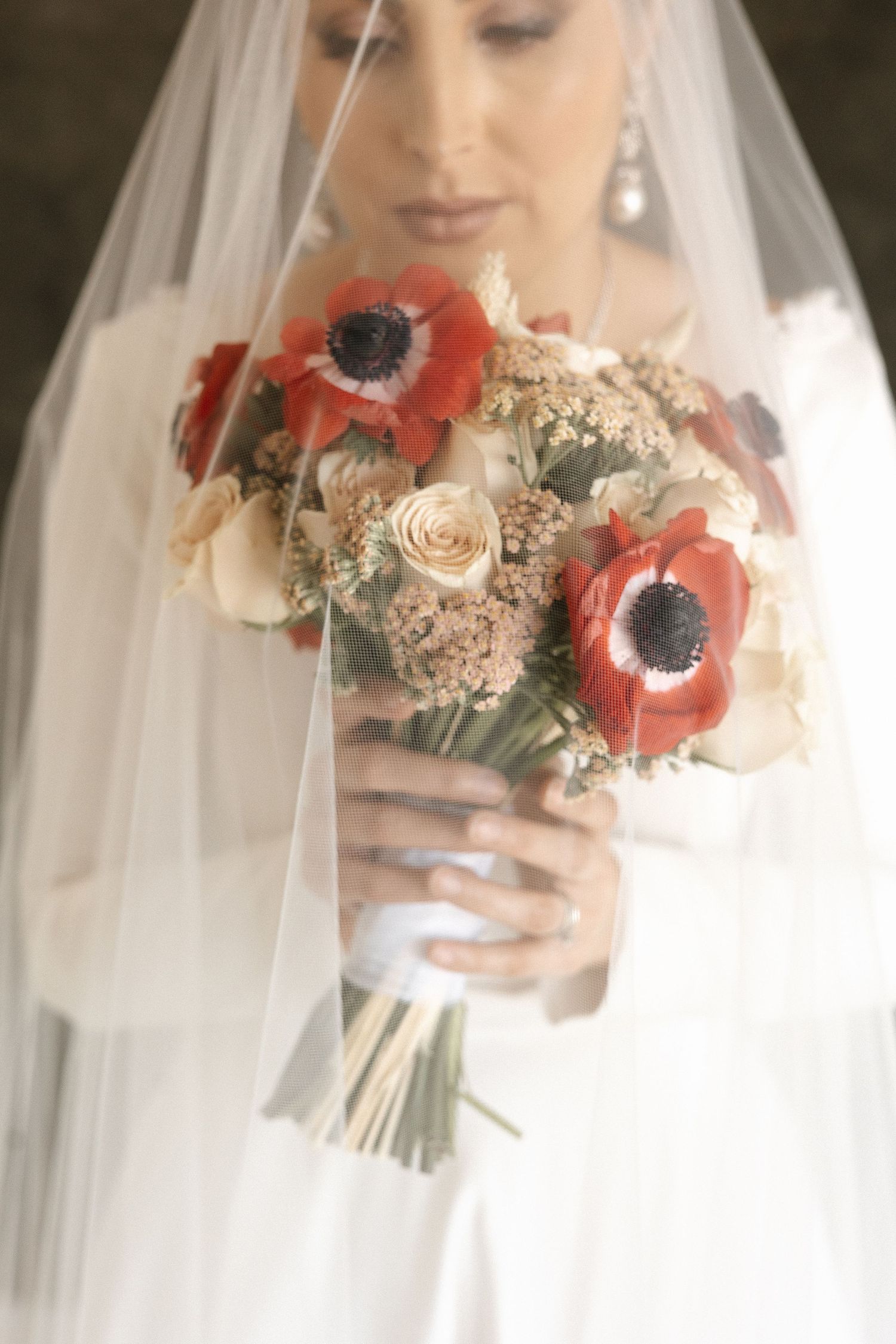 Close up of bridal bouquet with red anemones and cream roses under white veil.