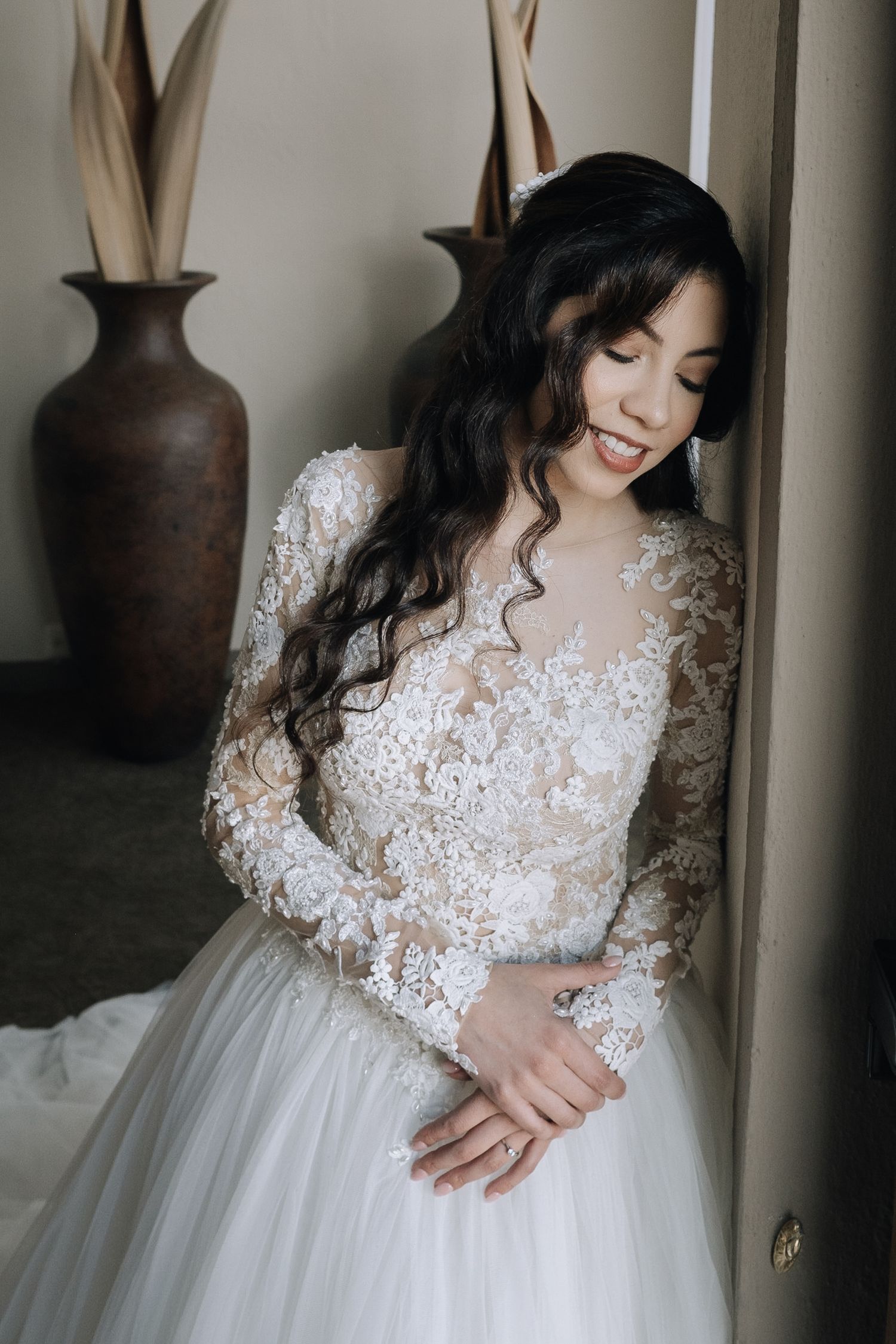 A bride in a delicate lace long sleeve wedding gown smiles softly while leaning against a wall.