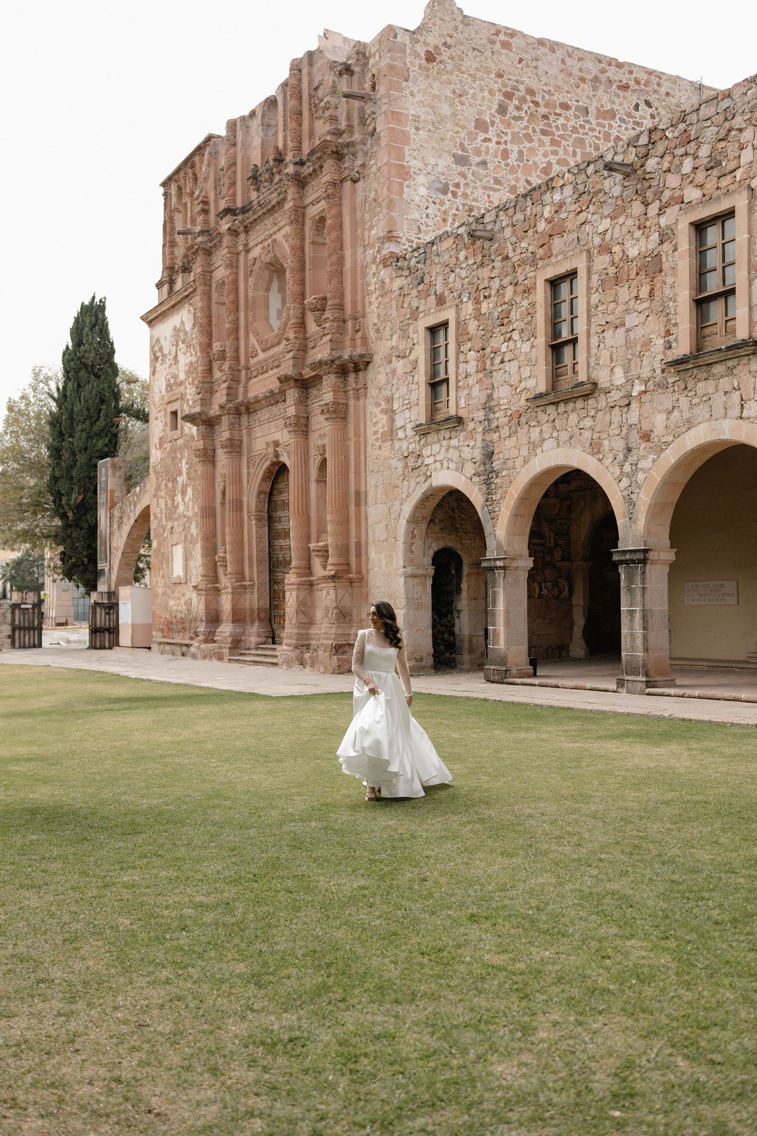 Stone archways of a historic mission building frame a grassy courtyard where a white wedding dress flows in the breeze.