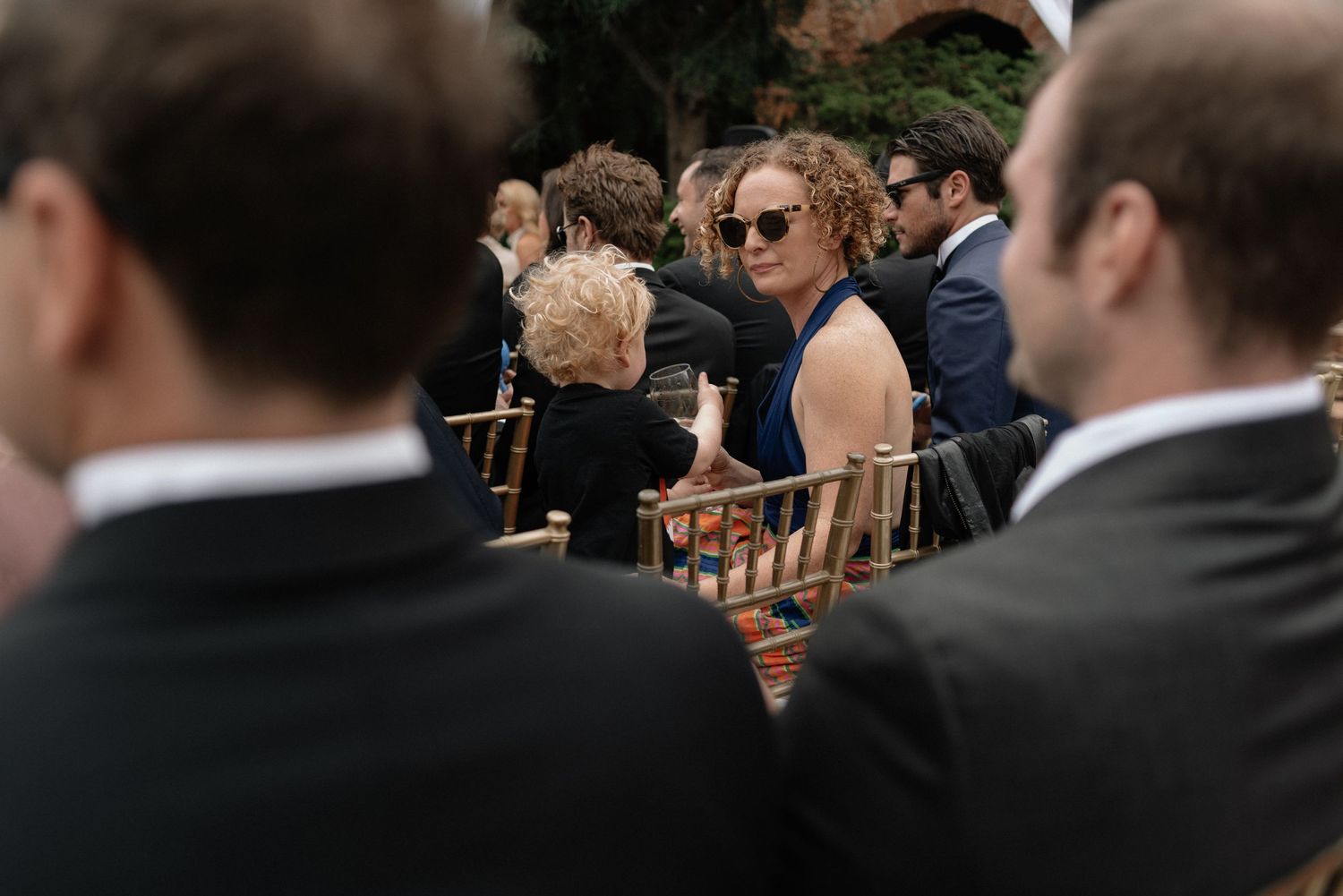 Guests seated at an outdoor event wearing formal attire with wooden chairs visible in the background.