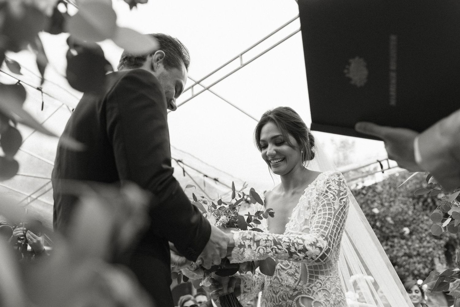 Black and white candid photo of a bride smiling during an outdoor garden wedding ceremony.