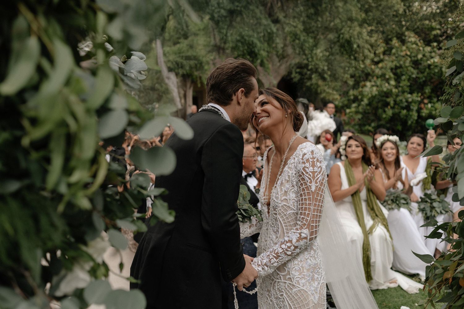 A romantic wedding ceremony moment outdoors with guests clapping in the background amid green foliage.