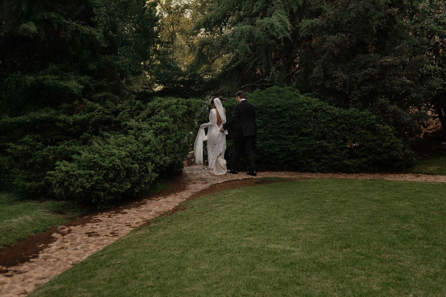 A couple in wedding attire walk along a garden path surrounded by lush greenery and manicured lawns.
