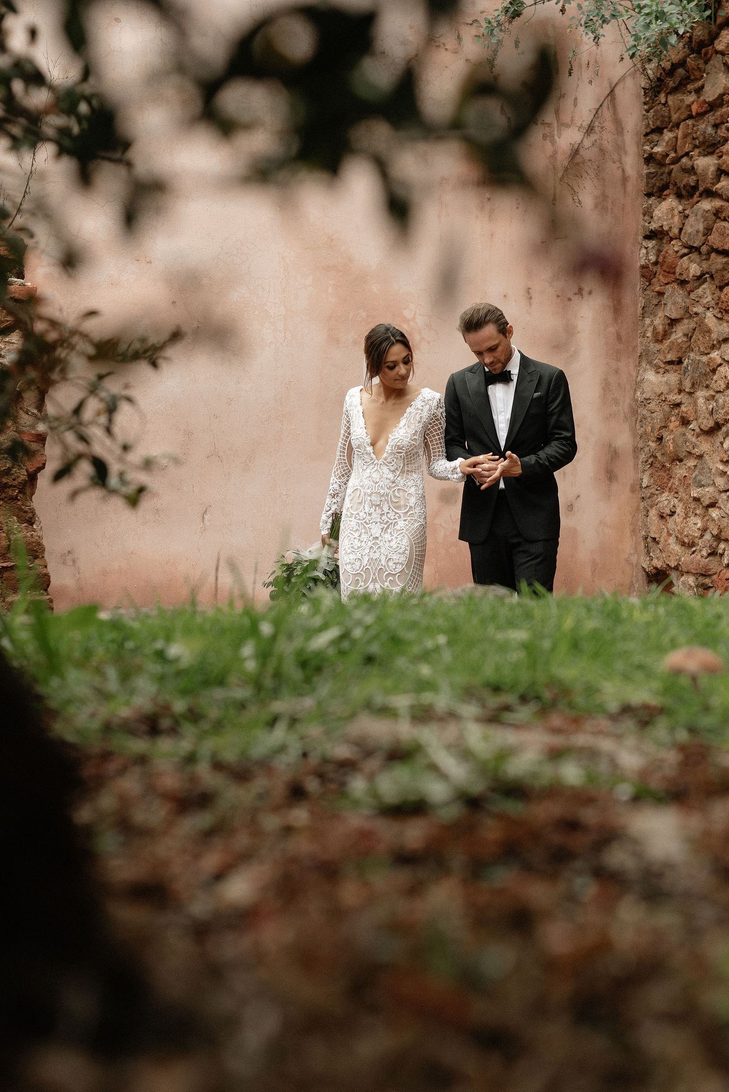 A couple in wedding attire stands together on a garden path framed by stone walls and greenery.