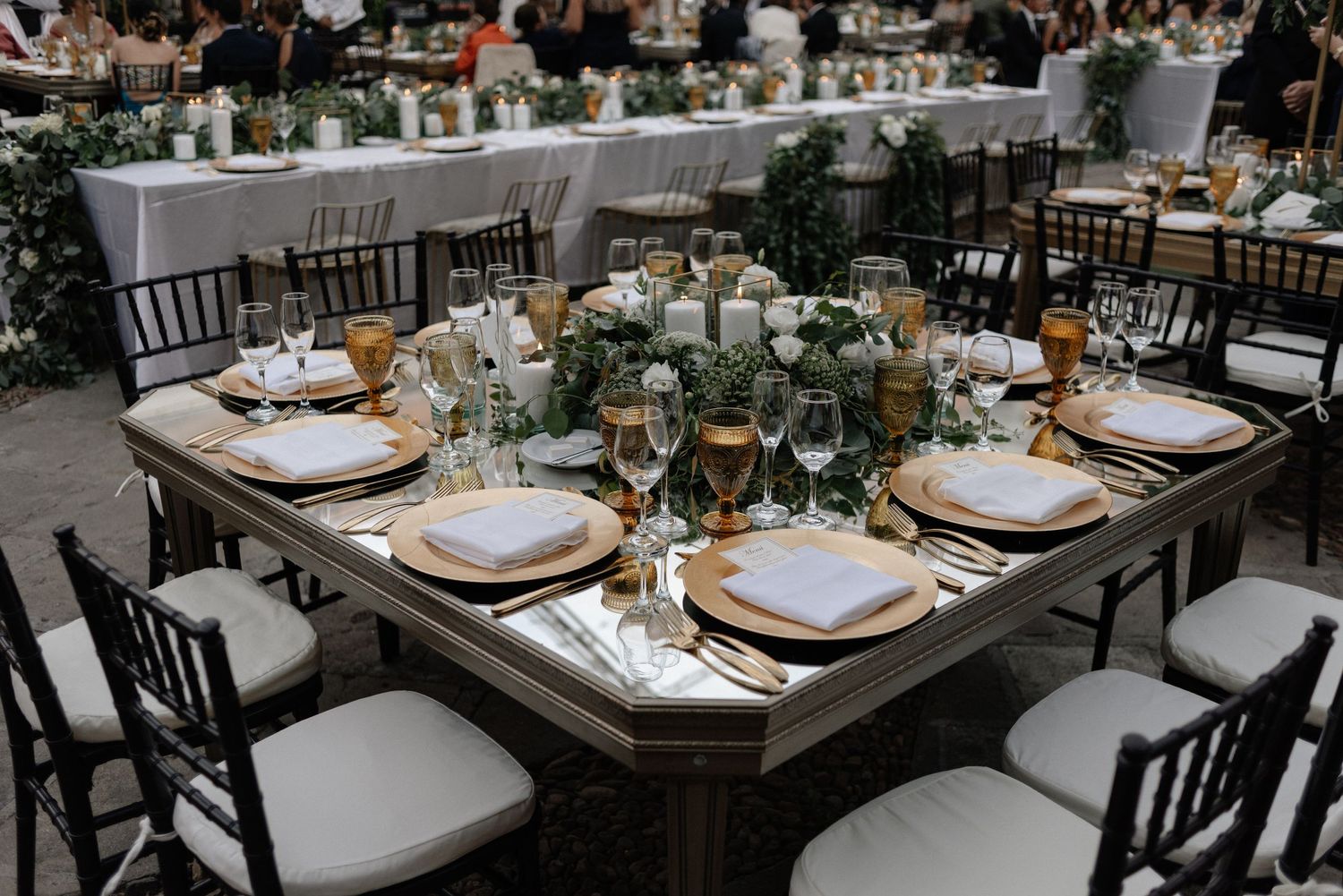 Elegant wedding reception table setting with white plates, dark wooden chairs and lush greenery centerpieces.