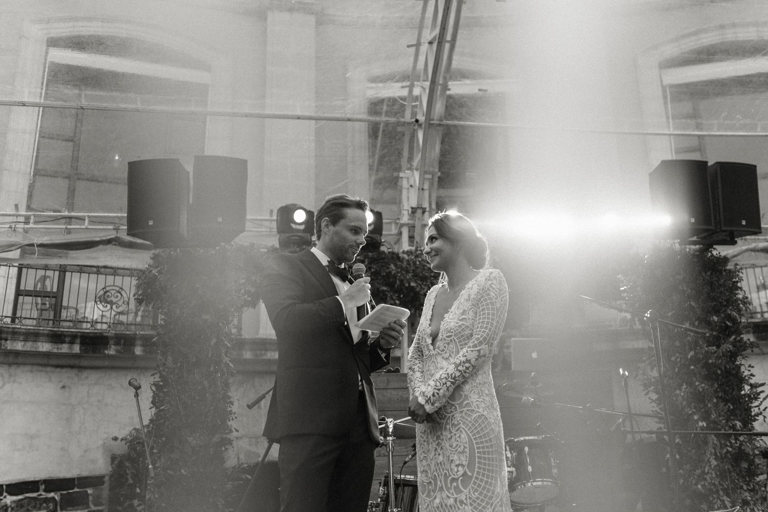 A romantic black and white wedding ceremony moment in front of an industrial backdrop with bright light shining behind.