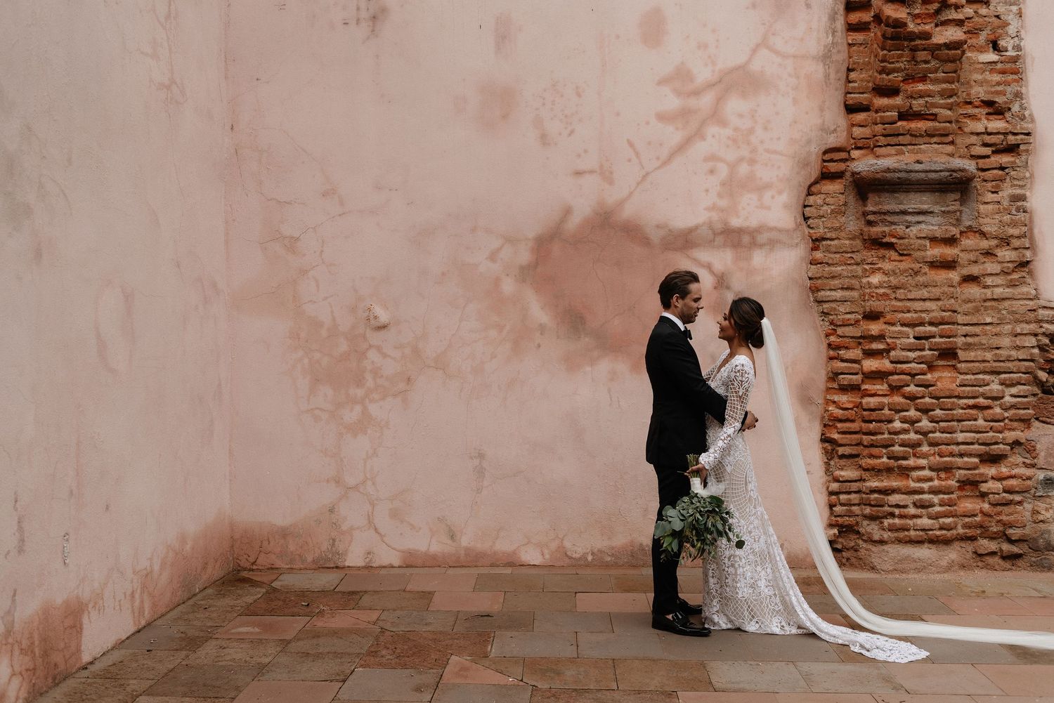 A wedding couple stands against a rustic pink wall and exposed brick at their romantic Mediterranean venue.