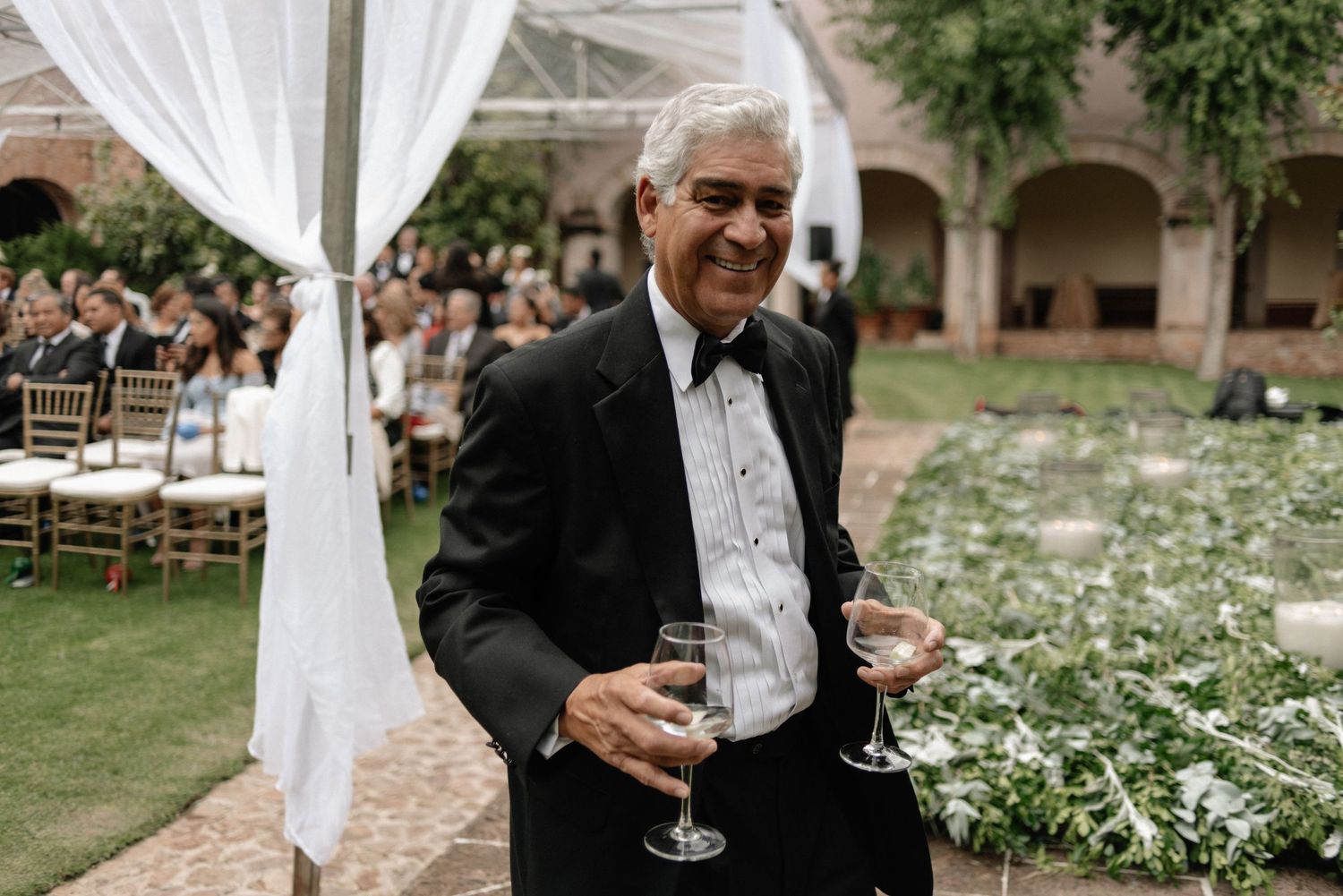A well-dressed guest in a dark suit smiles while holding a wine glass at an outdoor wedding reception.