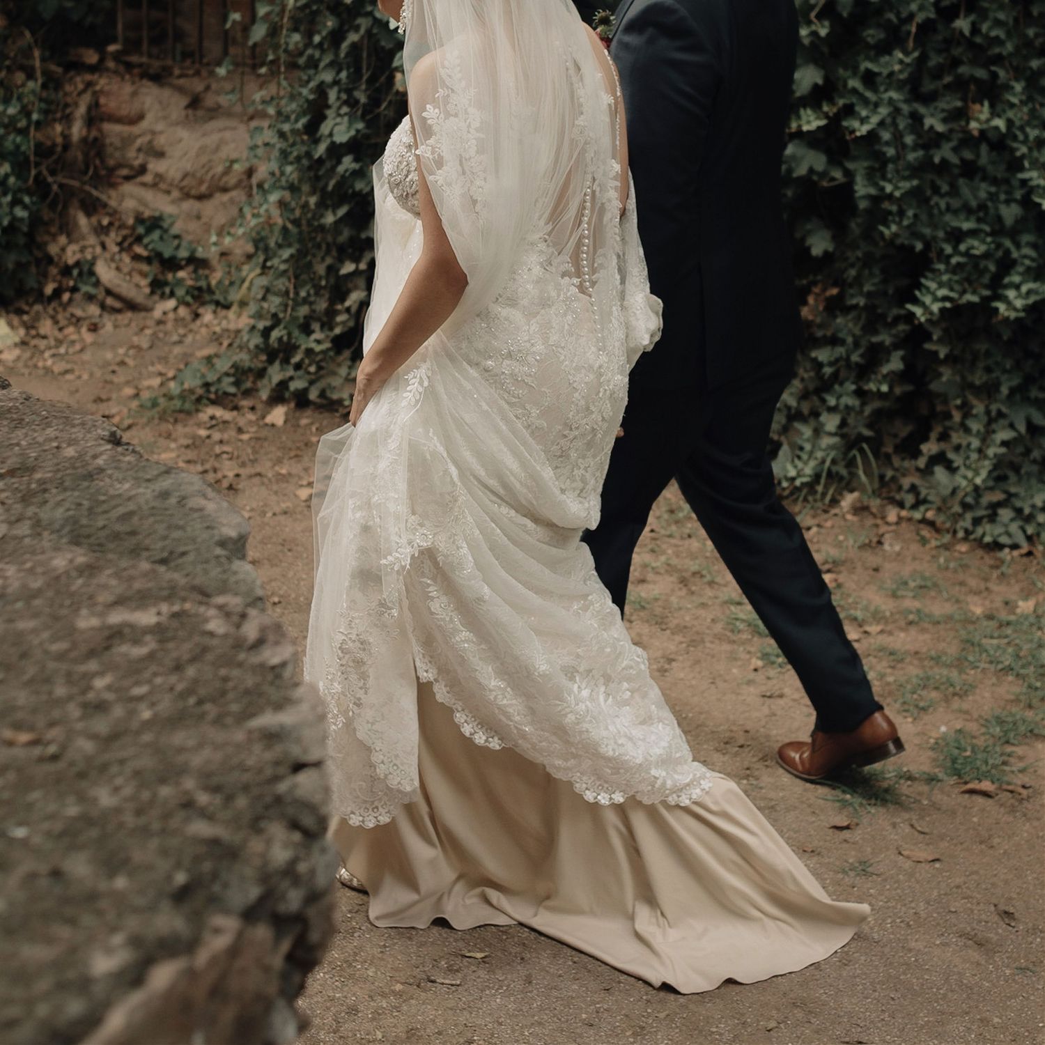 Close-up of bride's flowing lace wedding dress train and groom's black pants as they walk on a dirt path outdoors.