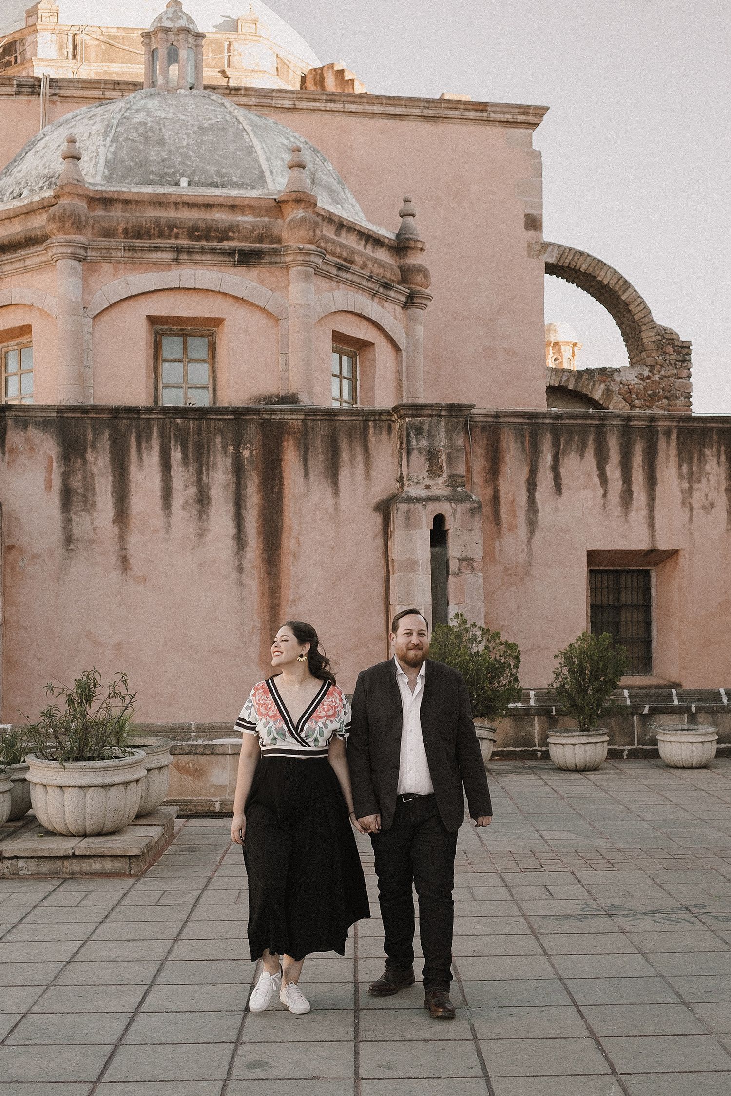 Couple walking and posing in front of a historic pink stone building with domed architecture.