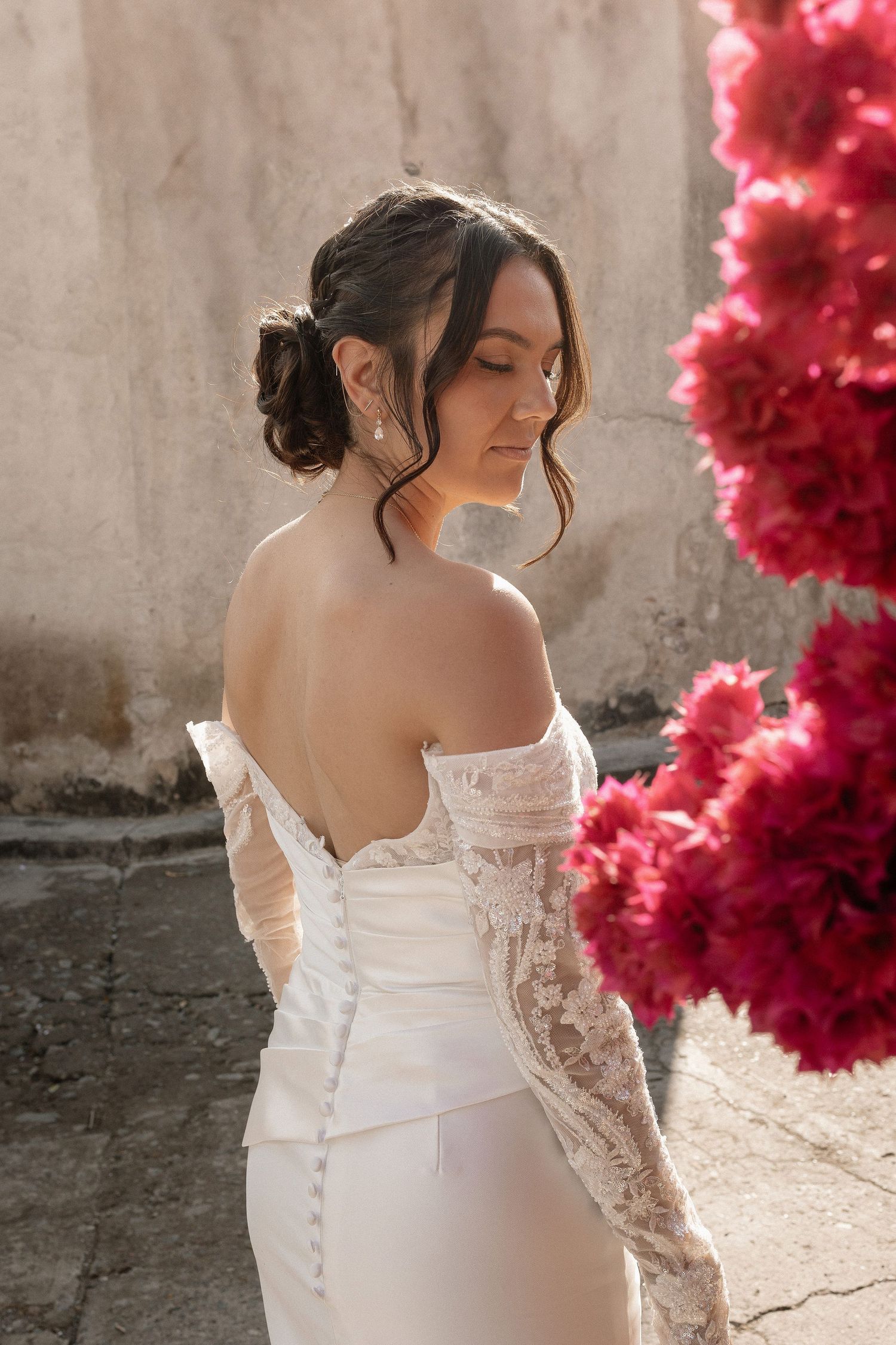 Close-up details of a white lace wedding dress with button closure against pink floral backdrop.
