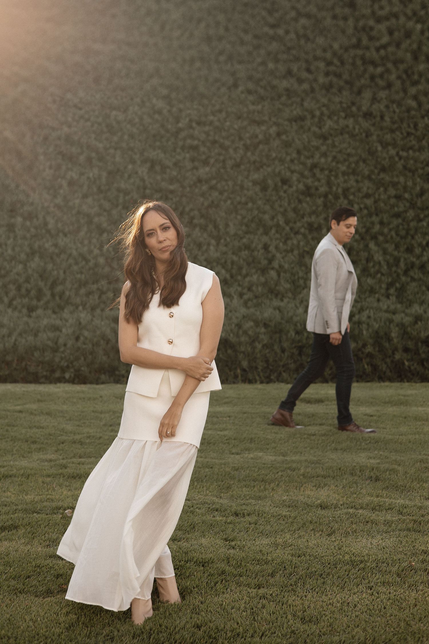 A woman in a white dress sits on grass while a figure walks in the background during golden hour.
