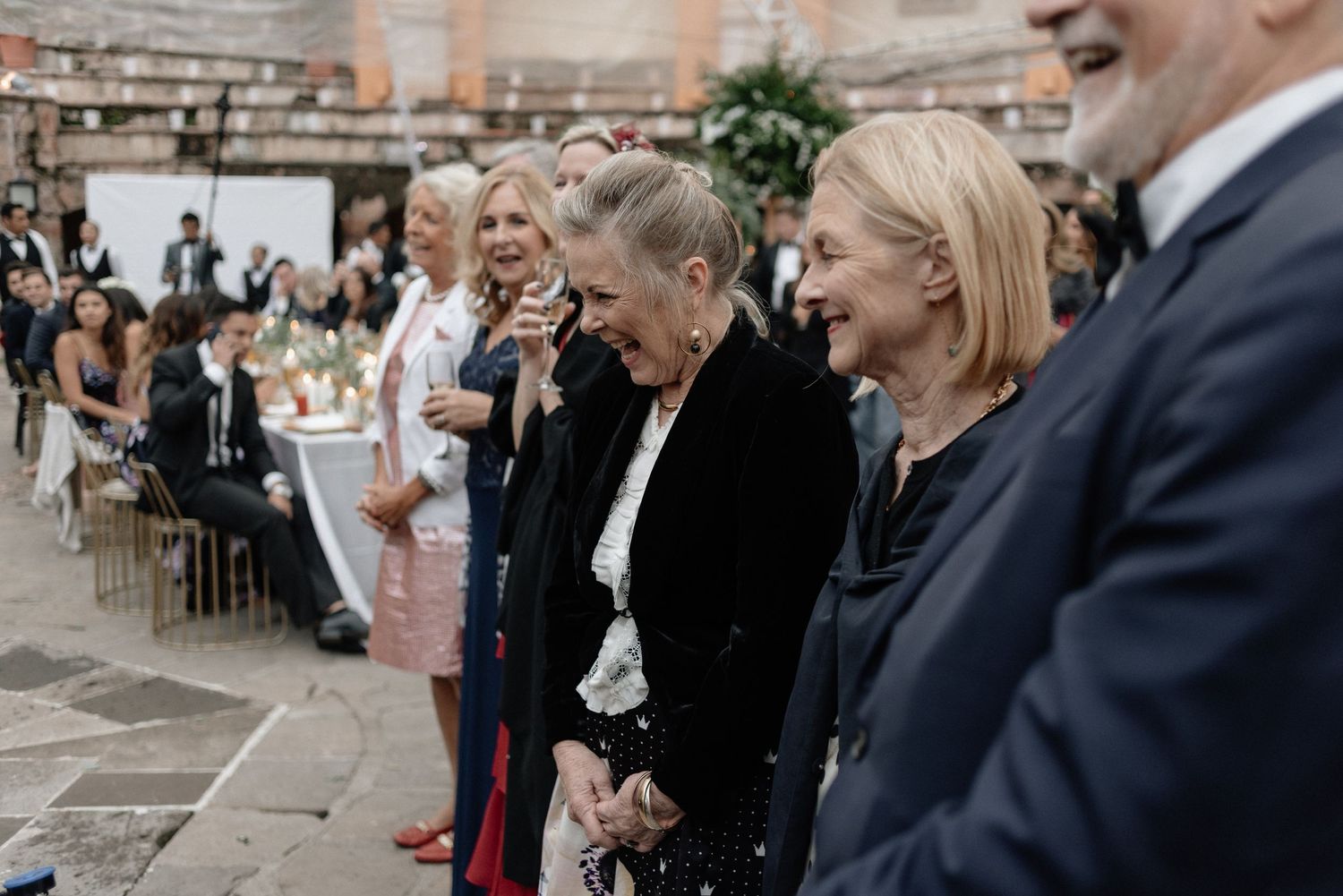 Wedding guests share joyful reactions during an outdoor ceremony at a rustic stone venue.
