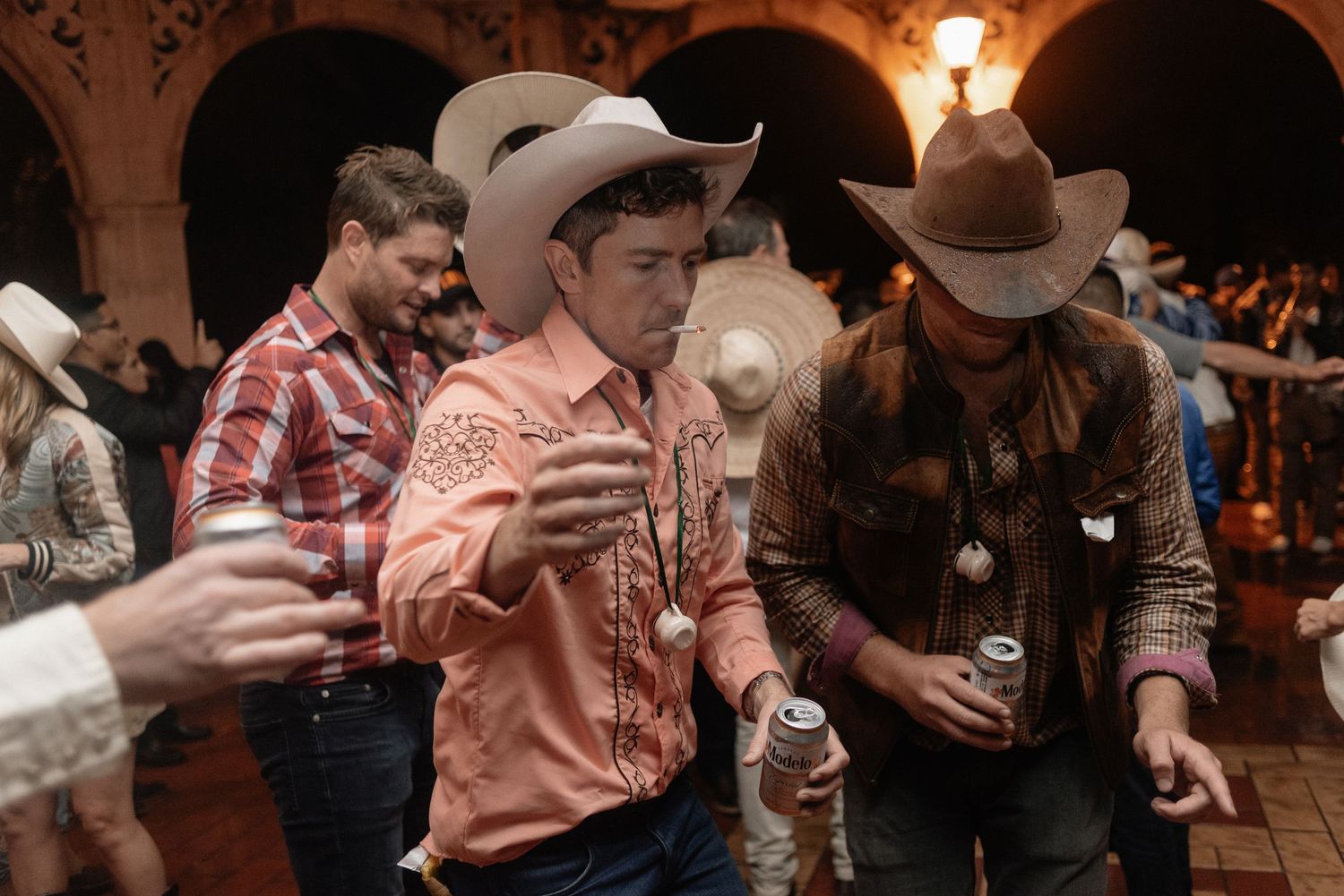 People in cowboy hats dancing and holding drinks at a western party in a rustic venue with warm lighting.