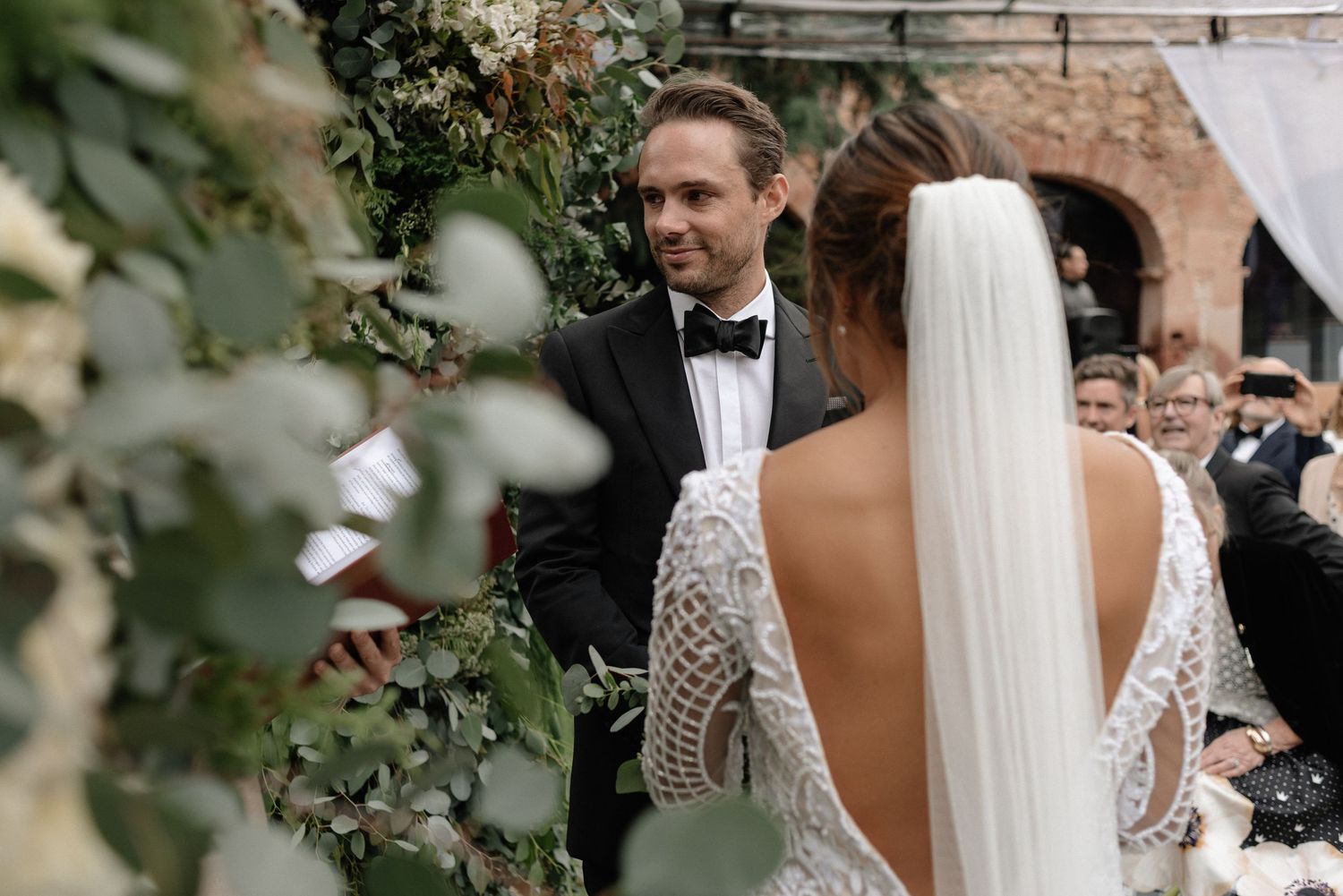 An elegant wedding ceremony outdoors features a couple exchanging vows among greenery and rustic brick walls.