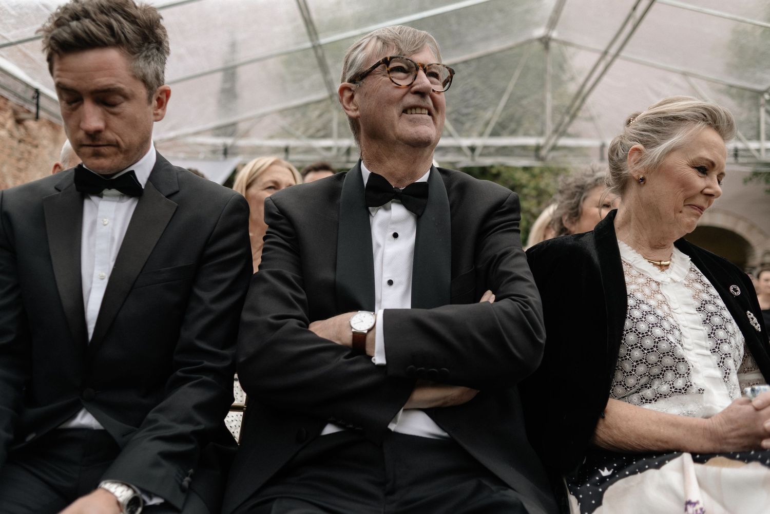 Three people in formal attire sit together at an outdoor event while appearing distant and unengaged.
