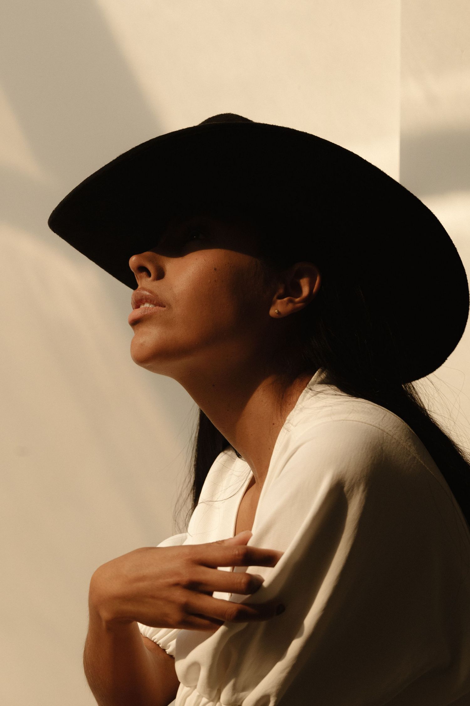 Woman in black wide-brim hat and white shirt poses gracefully against a light wall in warm natural light.