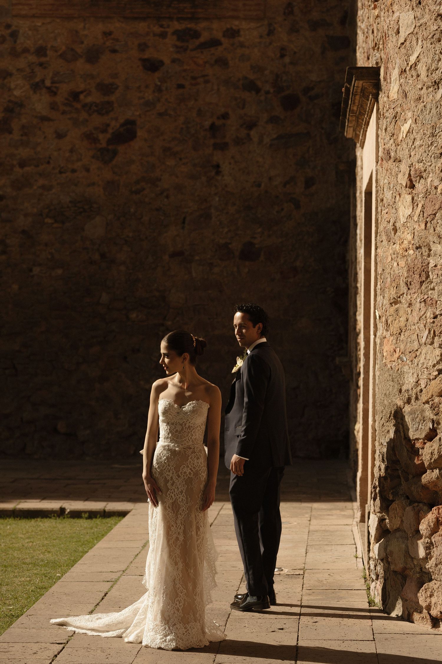 A couple in elegant wedding attire stands in warm sunlight against a rustic stone wall and courtyard.