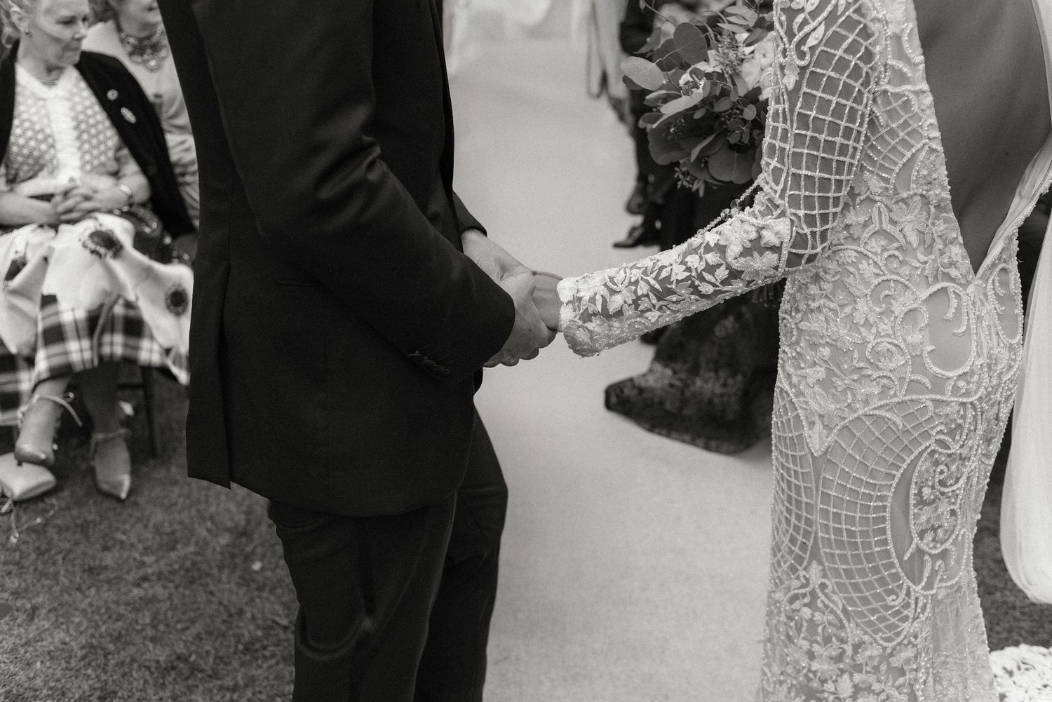 Black and white wedding photo showing close-up of couple holding hands during outdoor ceremony.