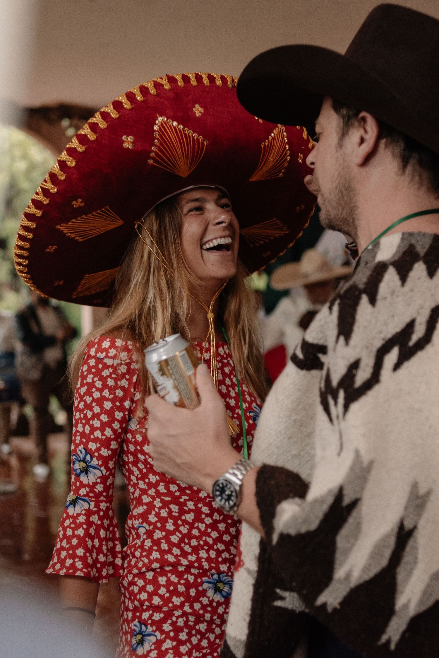 Person wearing red floral dress and sombrero laughs joyfully while holding a drink at festive celebration.