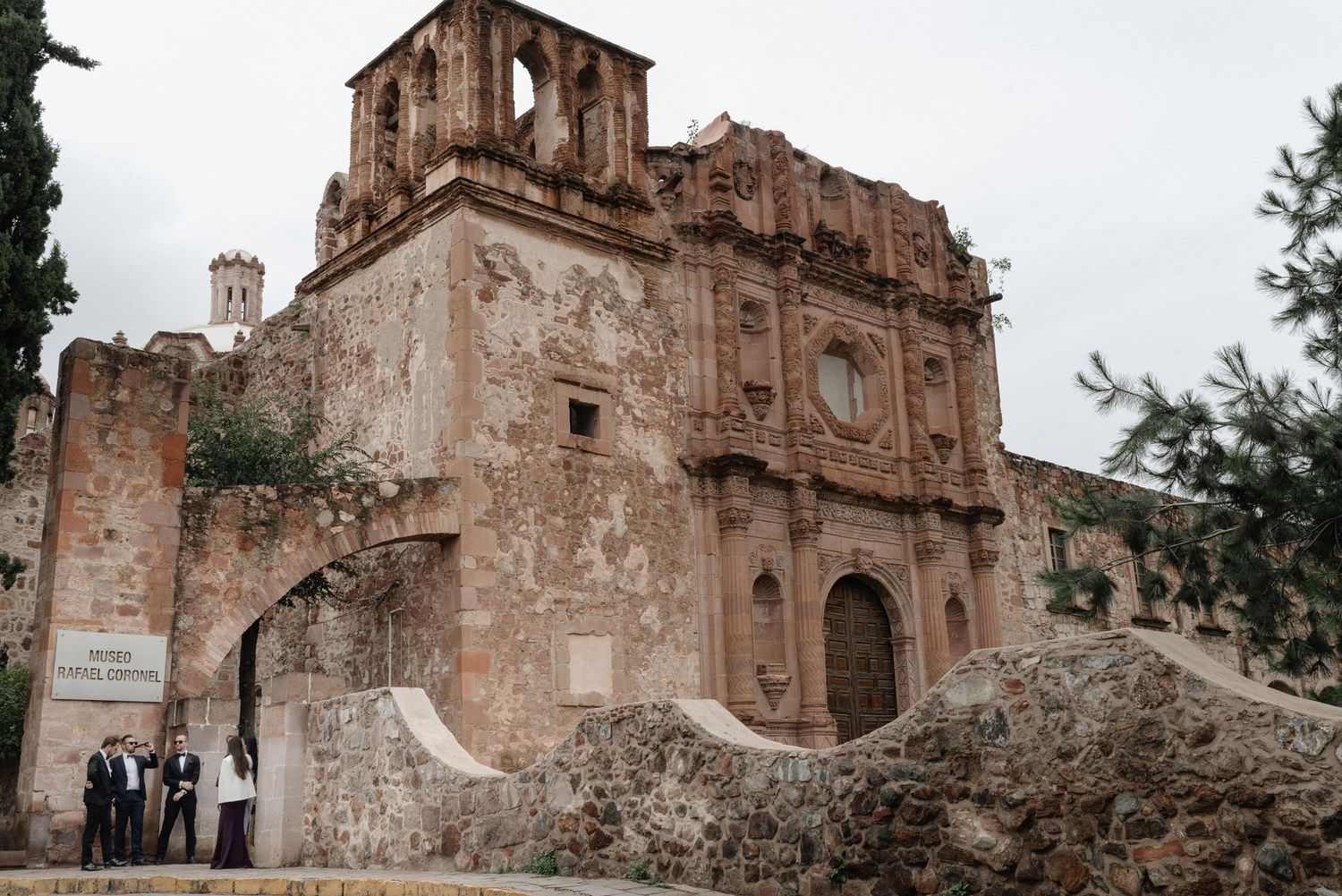 Ancient ruins of a weathered stone church with ornate facade and bell tower in faded pink hues stand against cloudy sky.