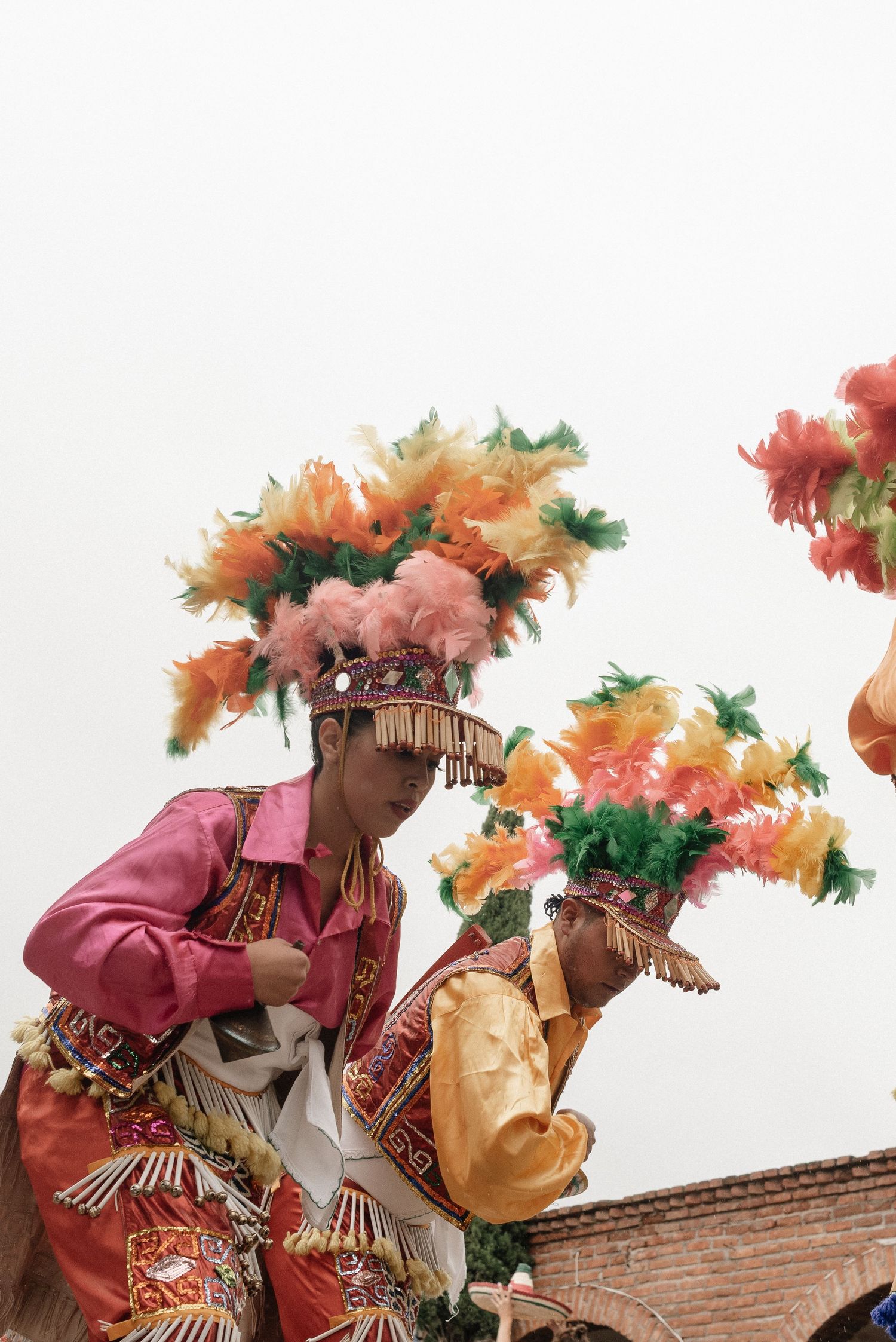 Dancers in colorful feathered headdresses and traditional costumes perform at festival near brick wall.