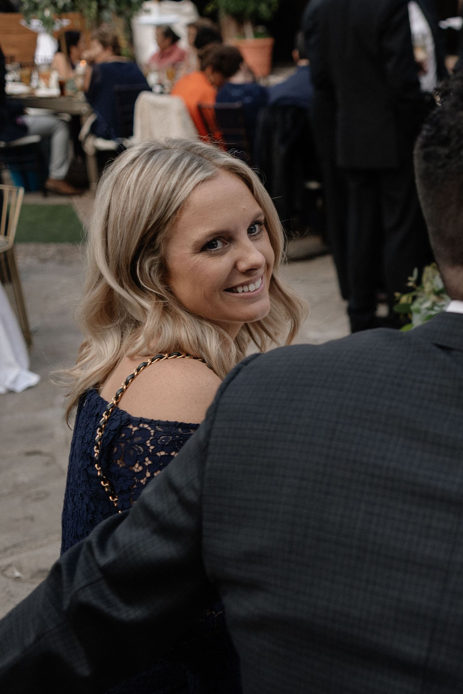 Woman in navy blue off-shoulder dress smiles at outdoor event with people mingling in background.