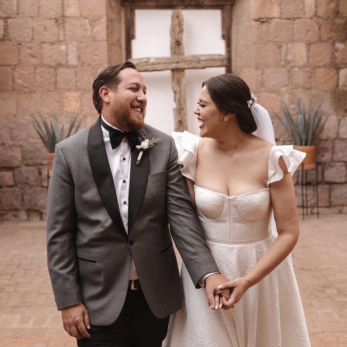 Wedding couple sharing a joyful moment in front of stone wall architecture with cross window.