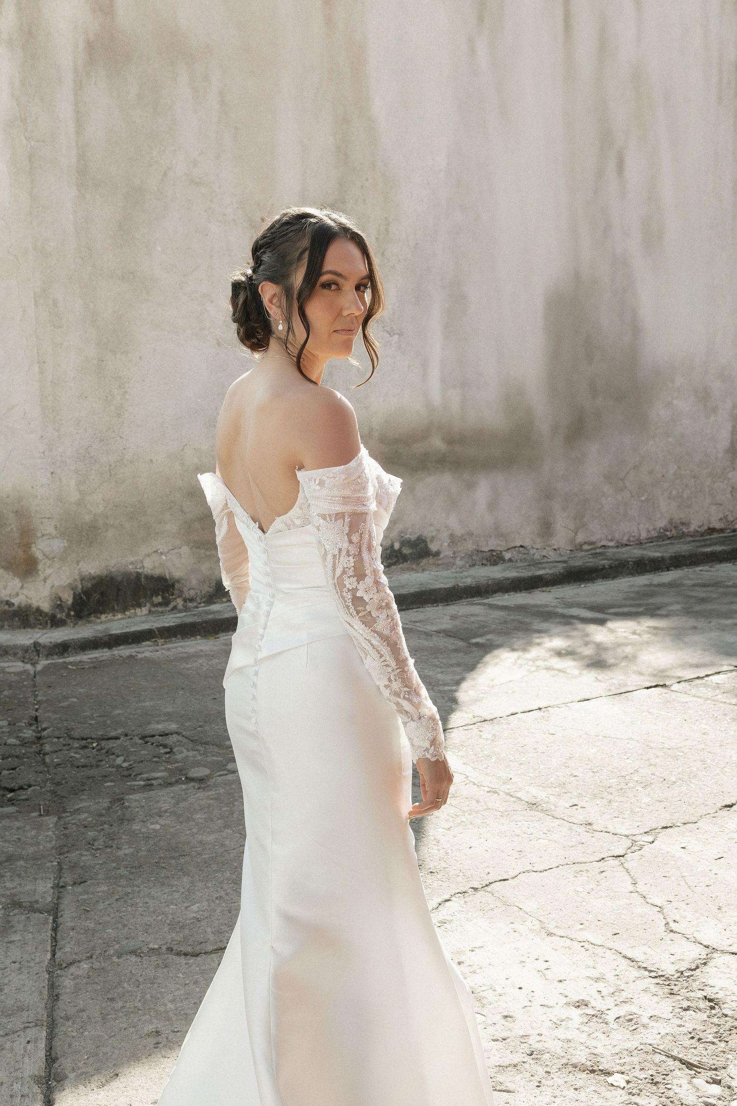 A bride in a flowing white lace wedding gown poses against a textured wall in natural lighting.