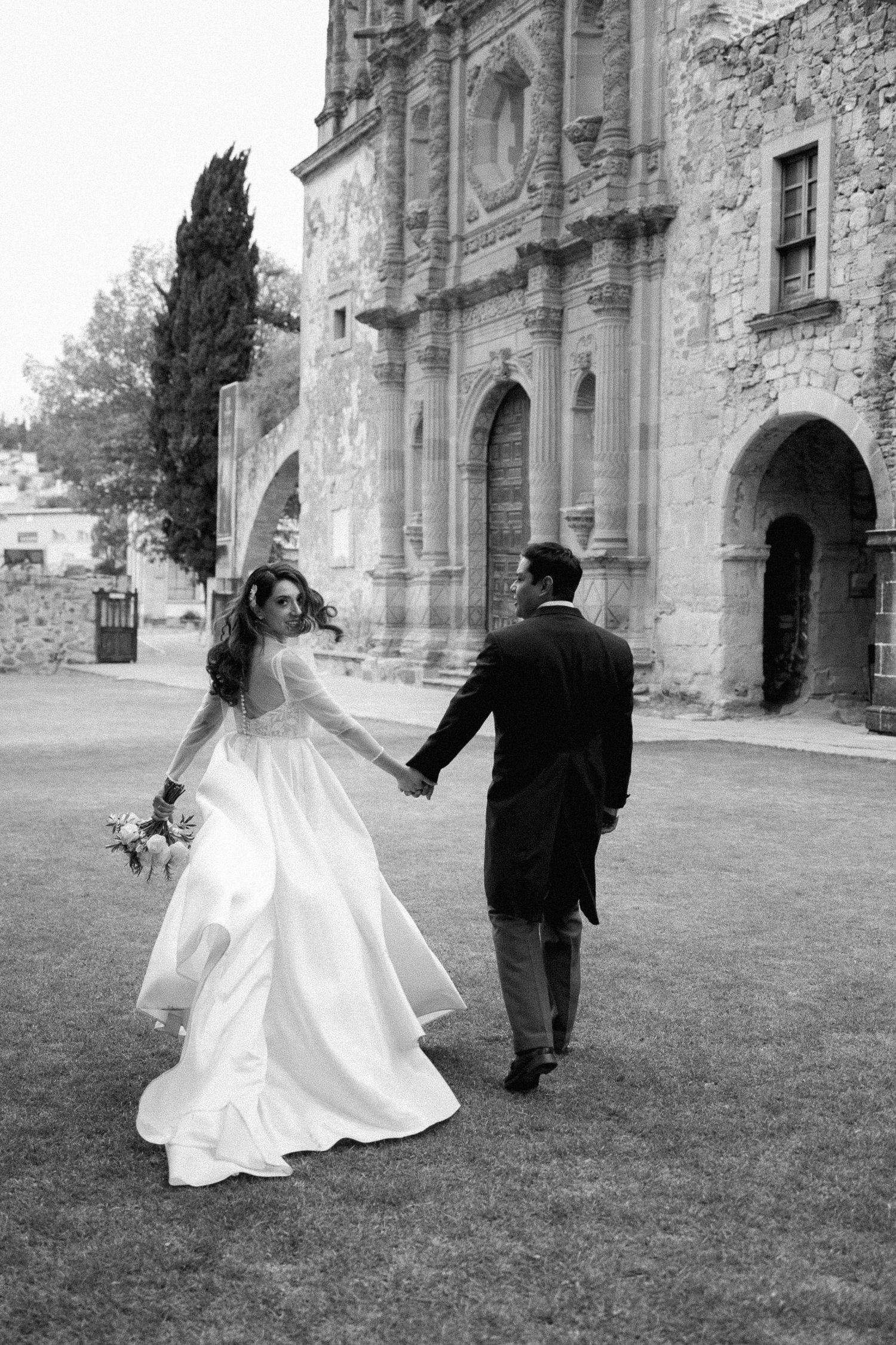 Newlyweds dance and twirl on grass in front of historic stone church architecture.