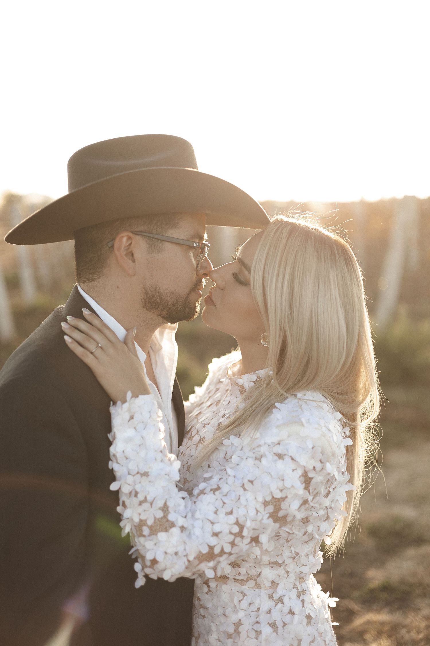 A romantic sunset photoshoot showing a couple in western attire embracing with the bride wearing a white floral dress.