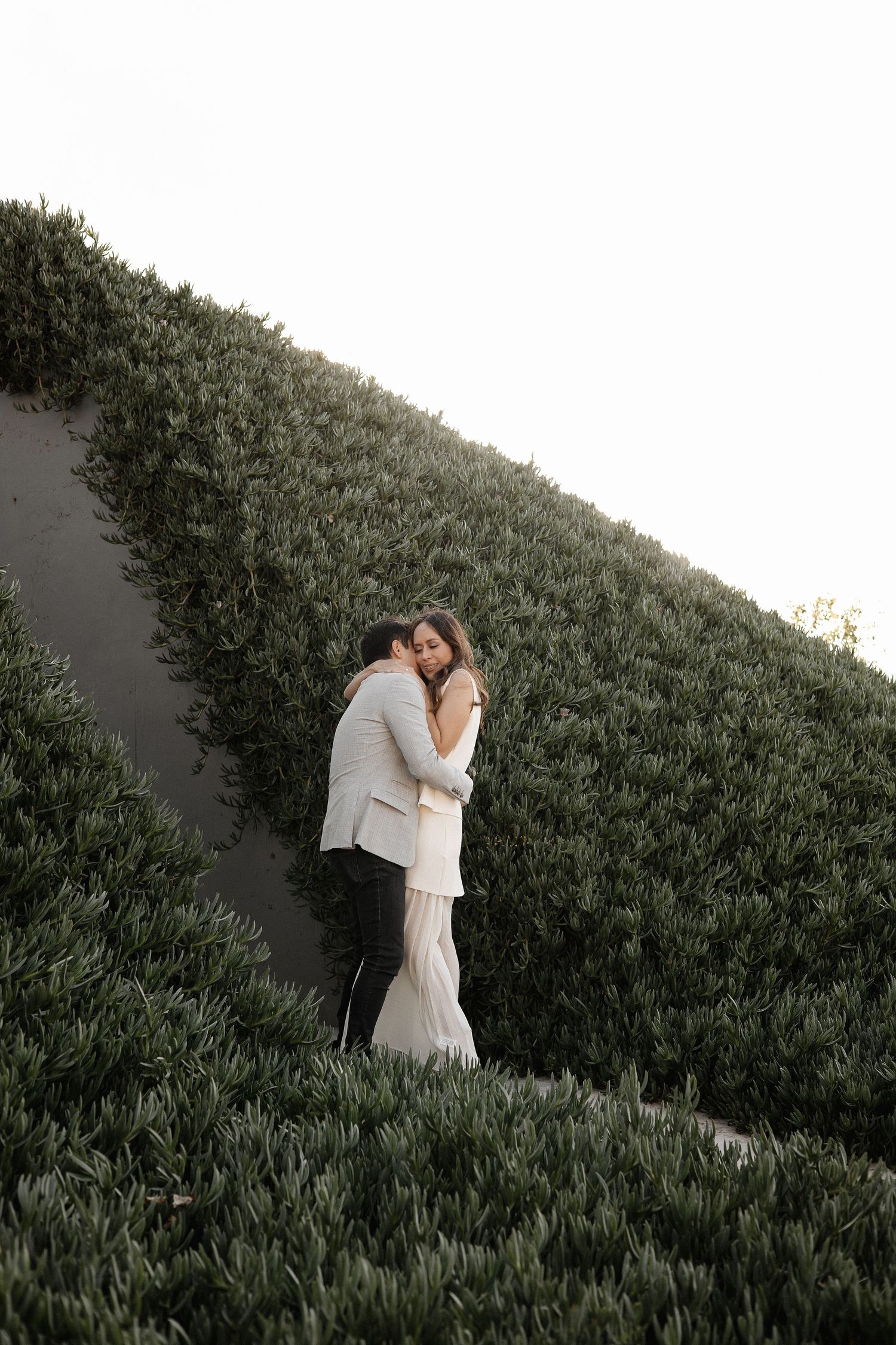 A couple in light colored outfits share an intimate embrace on a grassy hillside next to a hedge wall.