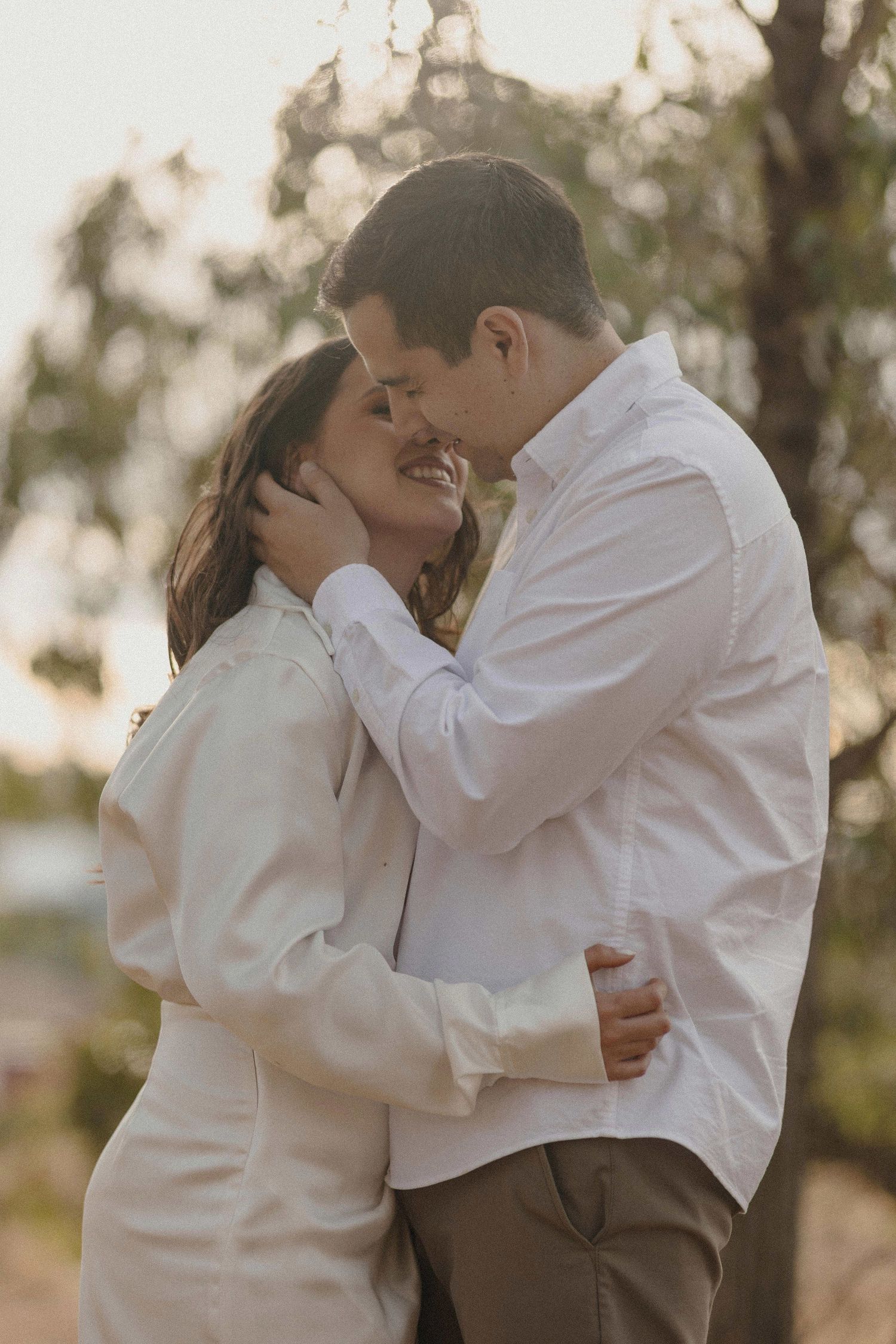 A romantic couple in white shirts embraces and shares tender moments during a sunset photoshoot outdoors.