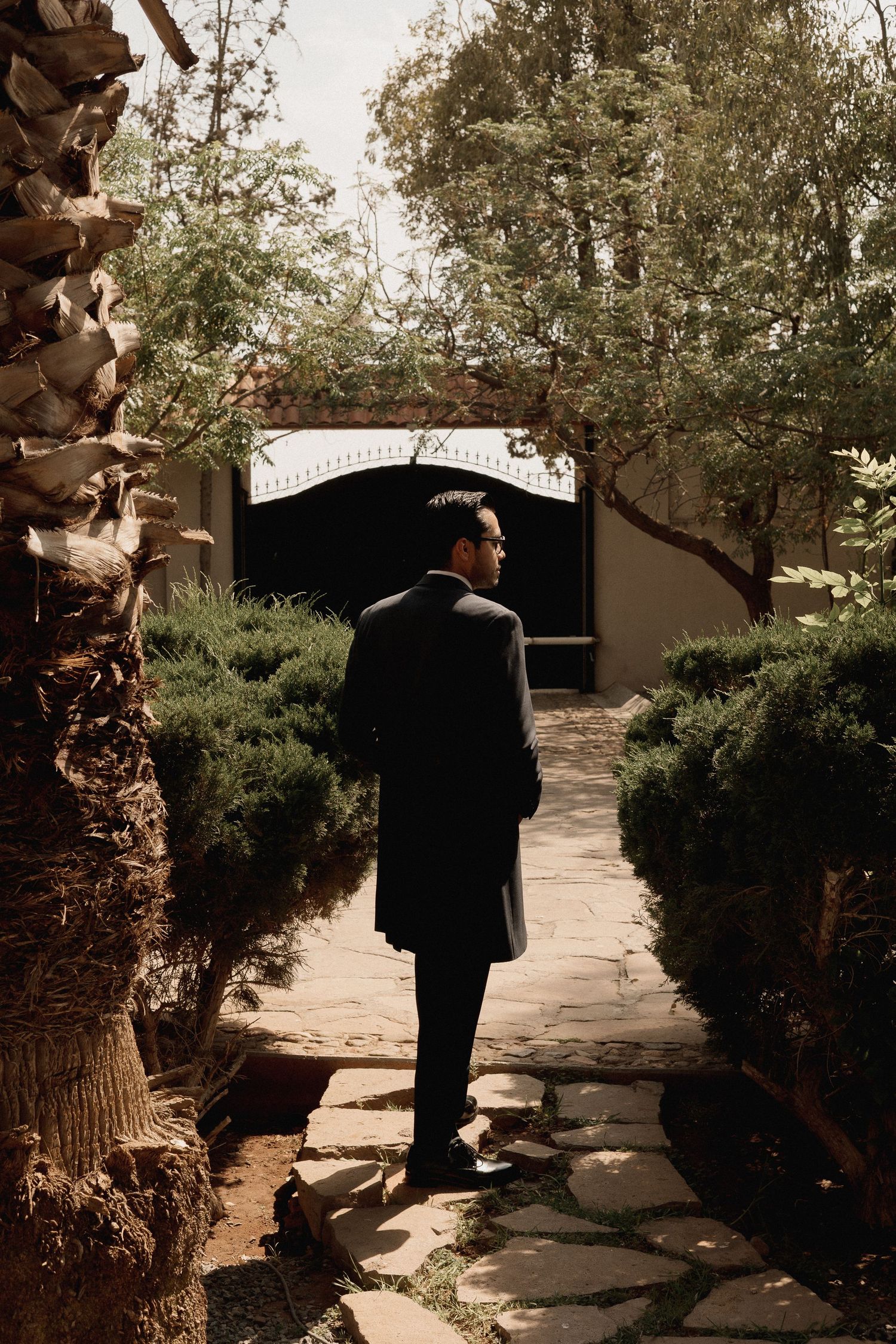 A person in a dark suit walks along a garden path between palm trees and bushes in dappled sunlight.