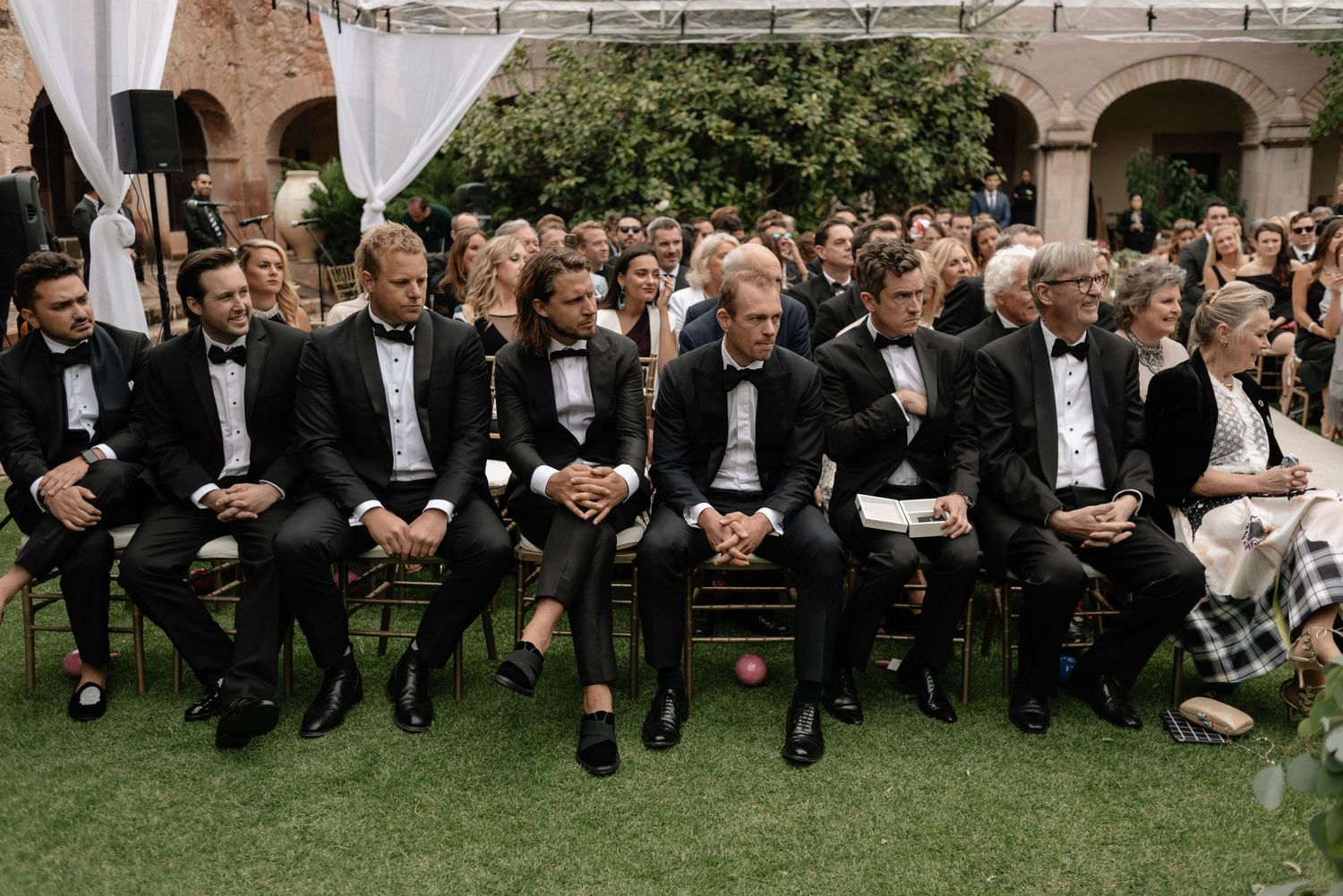 Wedding guests in black tuxedos seated at an elegant outdoor ceremony with stone arches in the background.