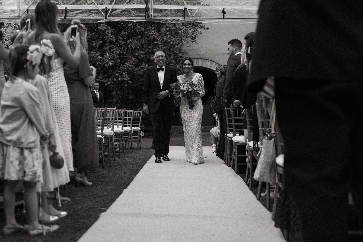 Vintage black and white photograph of an outdoor wedding ceremony with people lining a white aisle runner.