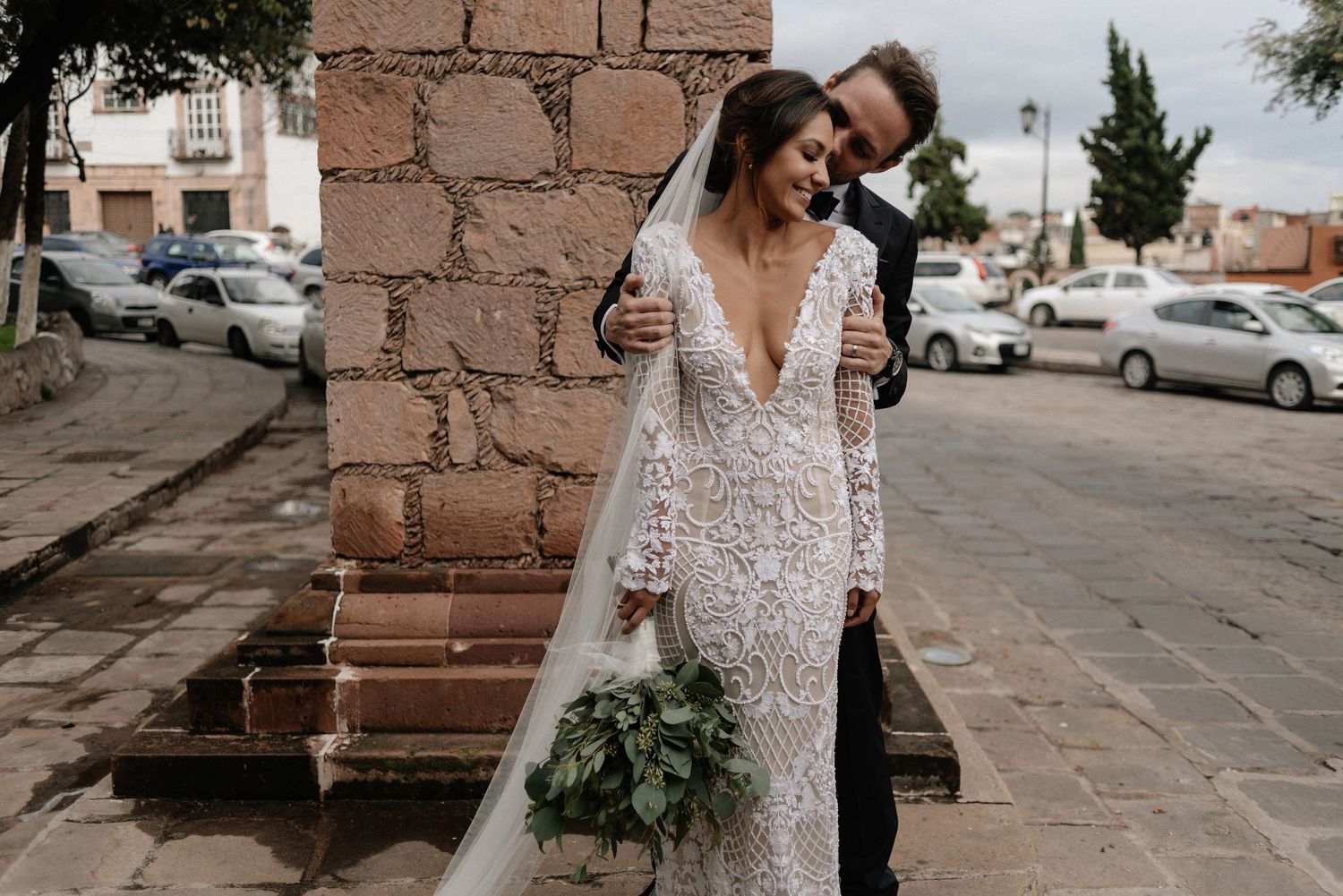 A romantic wedding couple stands against a rustic brick wall, the bride wearing a delicate lace gown holding a greenery bouquet.