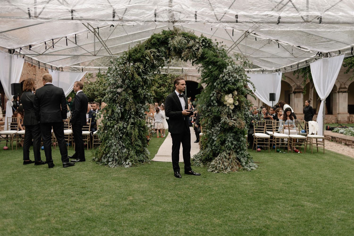 A lush green floral arch stands on grass at an elegant outdoor wedding reception under a white tent canopy.