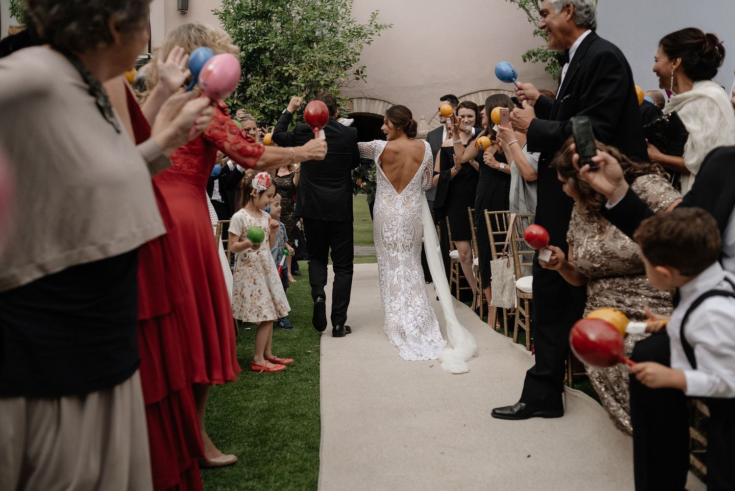 Wedding guests line a garden walkway with cameras and phones to capture a couple during their outdoor ceremony.
