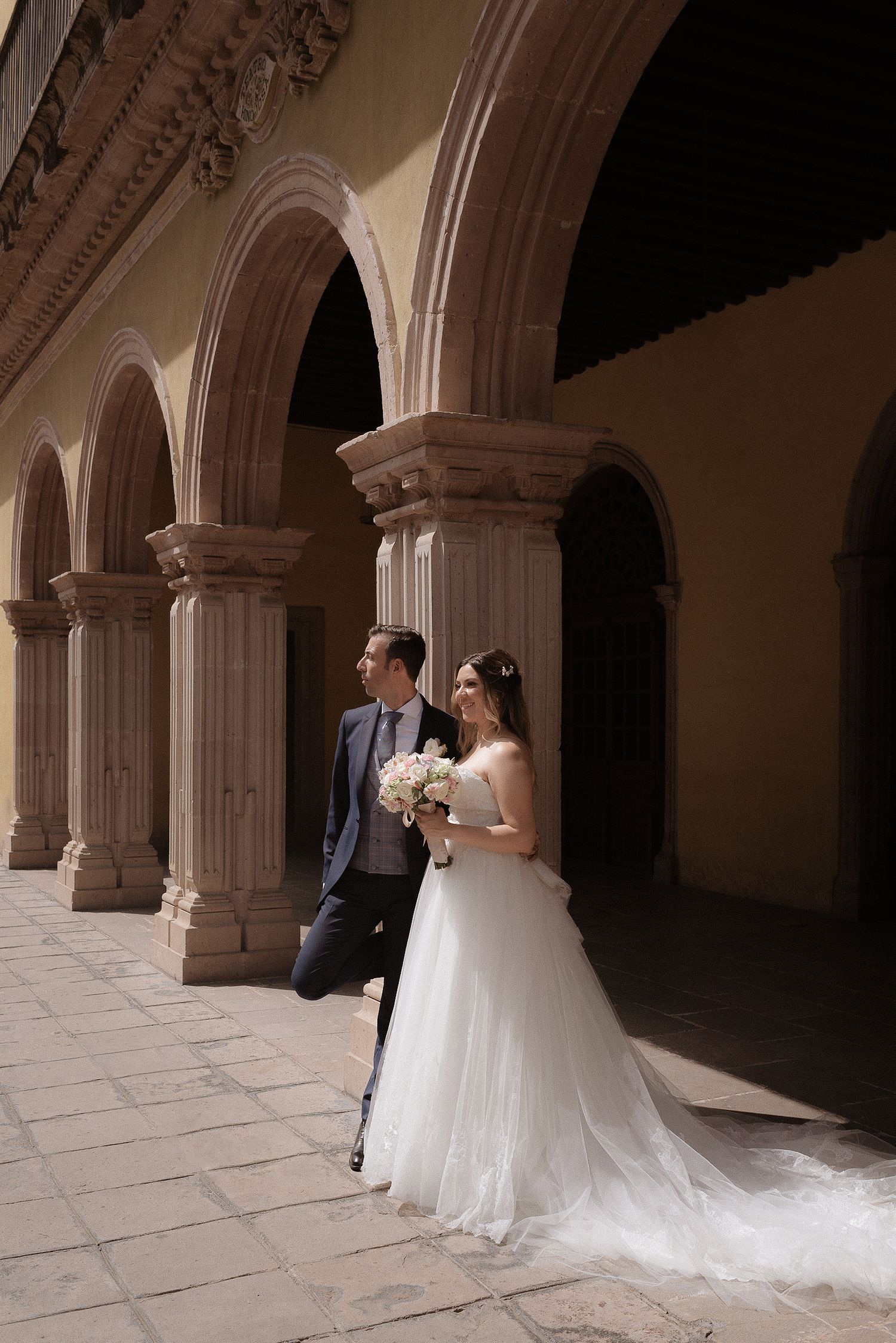 Wedding couple posing under ornate stone archways in a historic corridor.