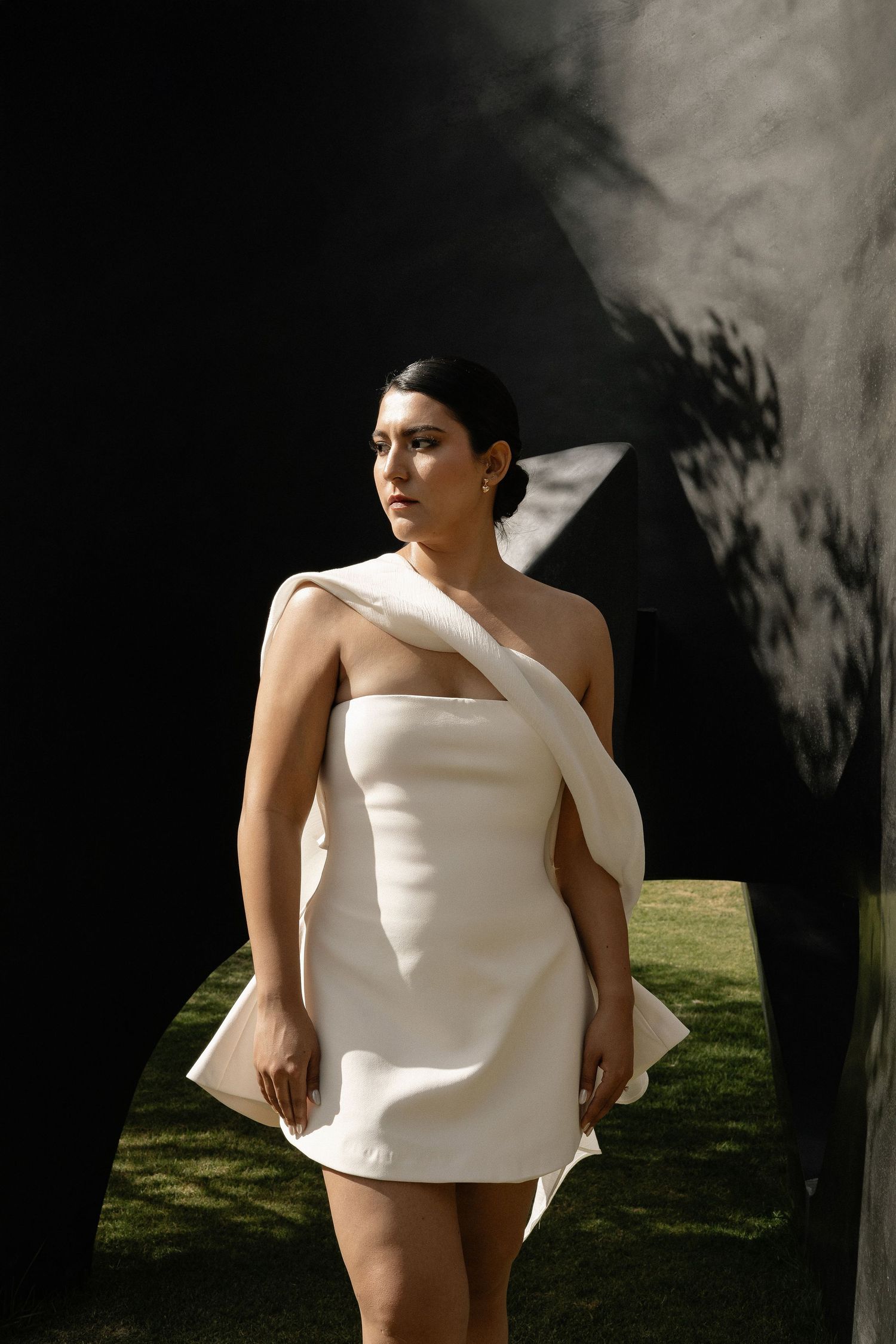 A figure in a sculptural white dress poses against a dark background with dappled light and shadows on grass.