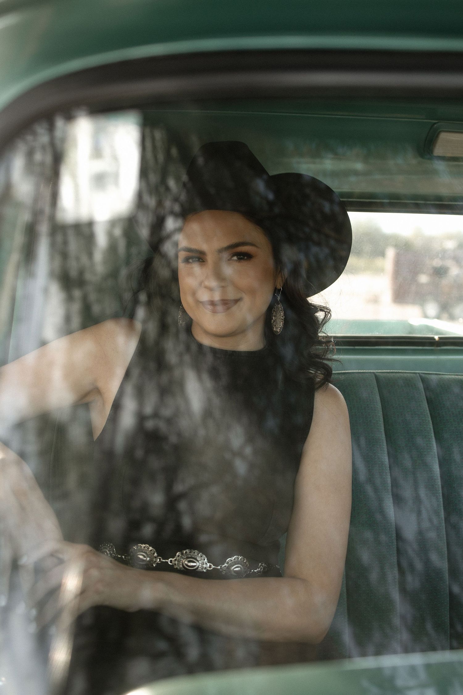 Woman in black dress sits in vintage car interior viewed through window reflections and shadows.