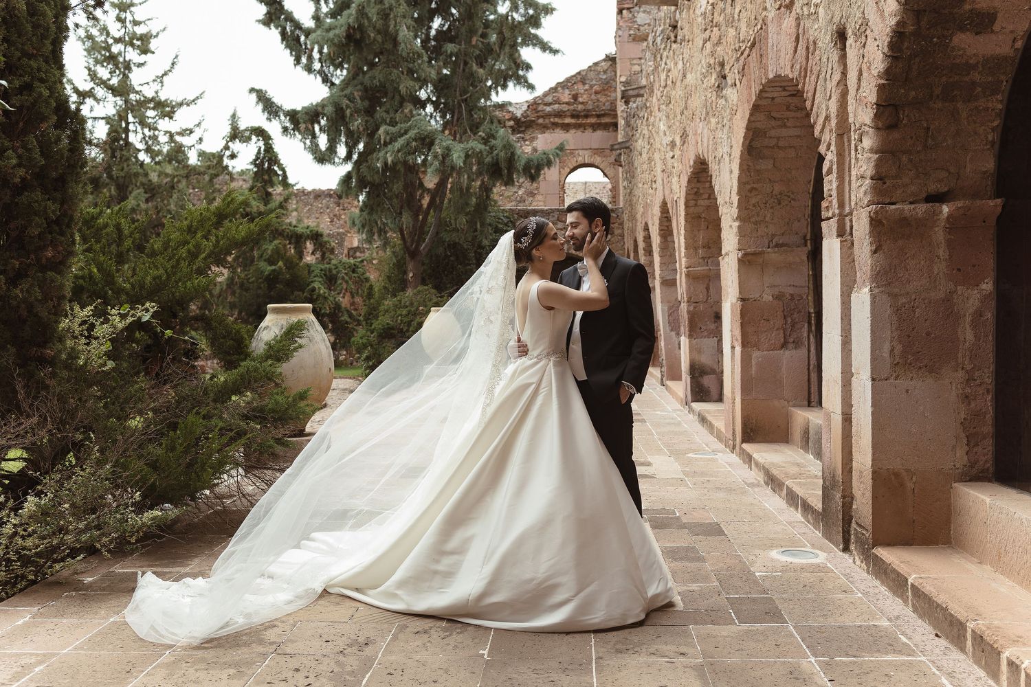A romantic wedding couple poses in an elegant stone archway corridor with a flowing white dress trailing behind them.