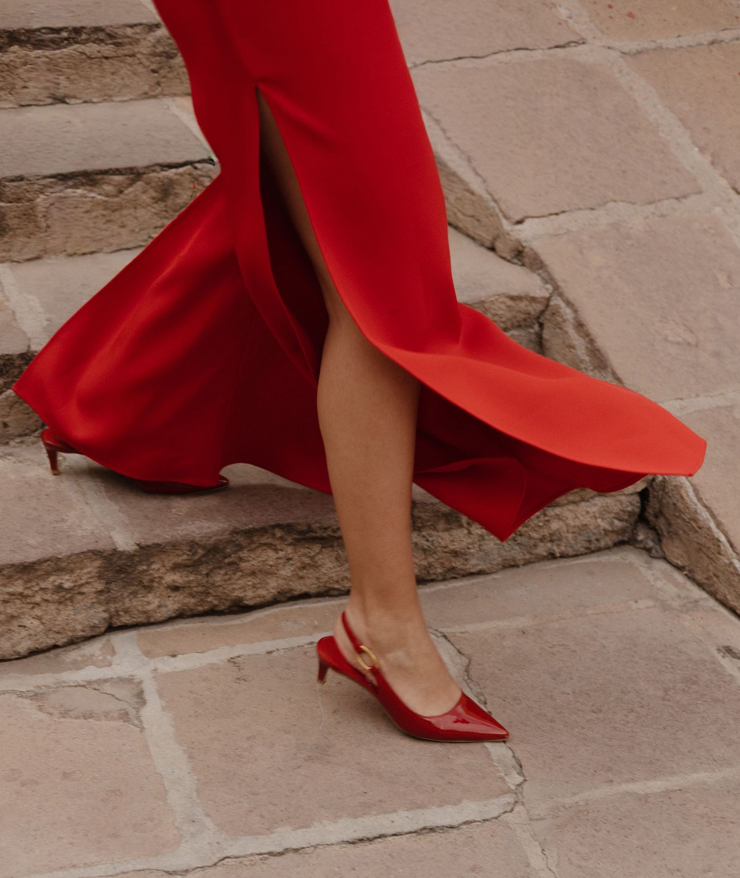 A person in red high heels walks up stone steps, their flowing red dress trailing elegantly behind them.
