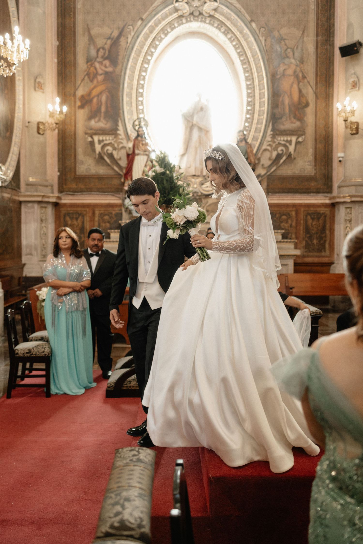 A wedding ceremony takes place in an ornate church with a red carpet aisle and elegant chandeliers.
