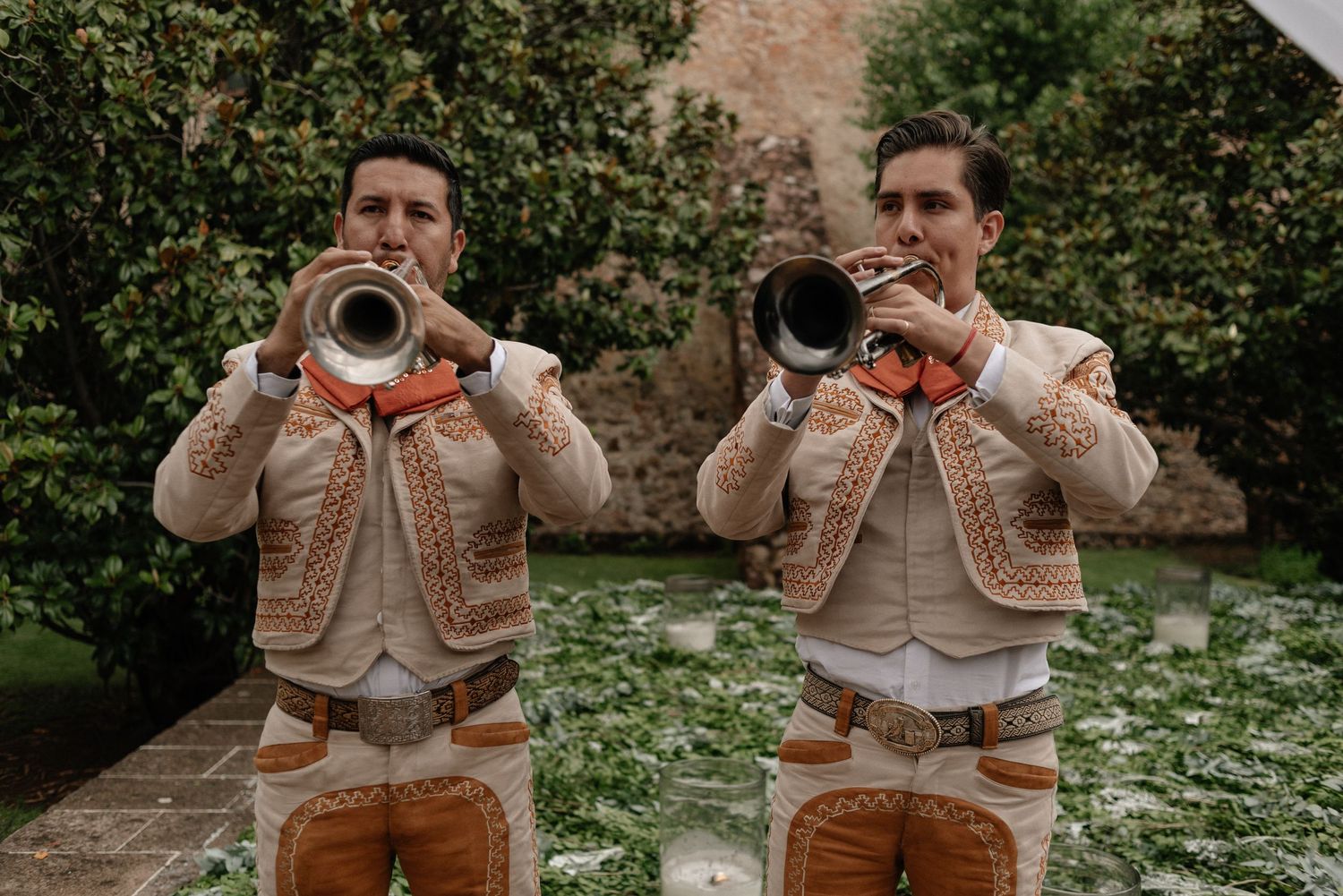 Two musicians in matching beige and brown outfits play trumpets outdoors in a garden setting.