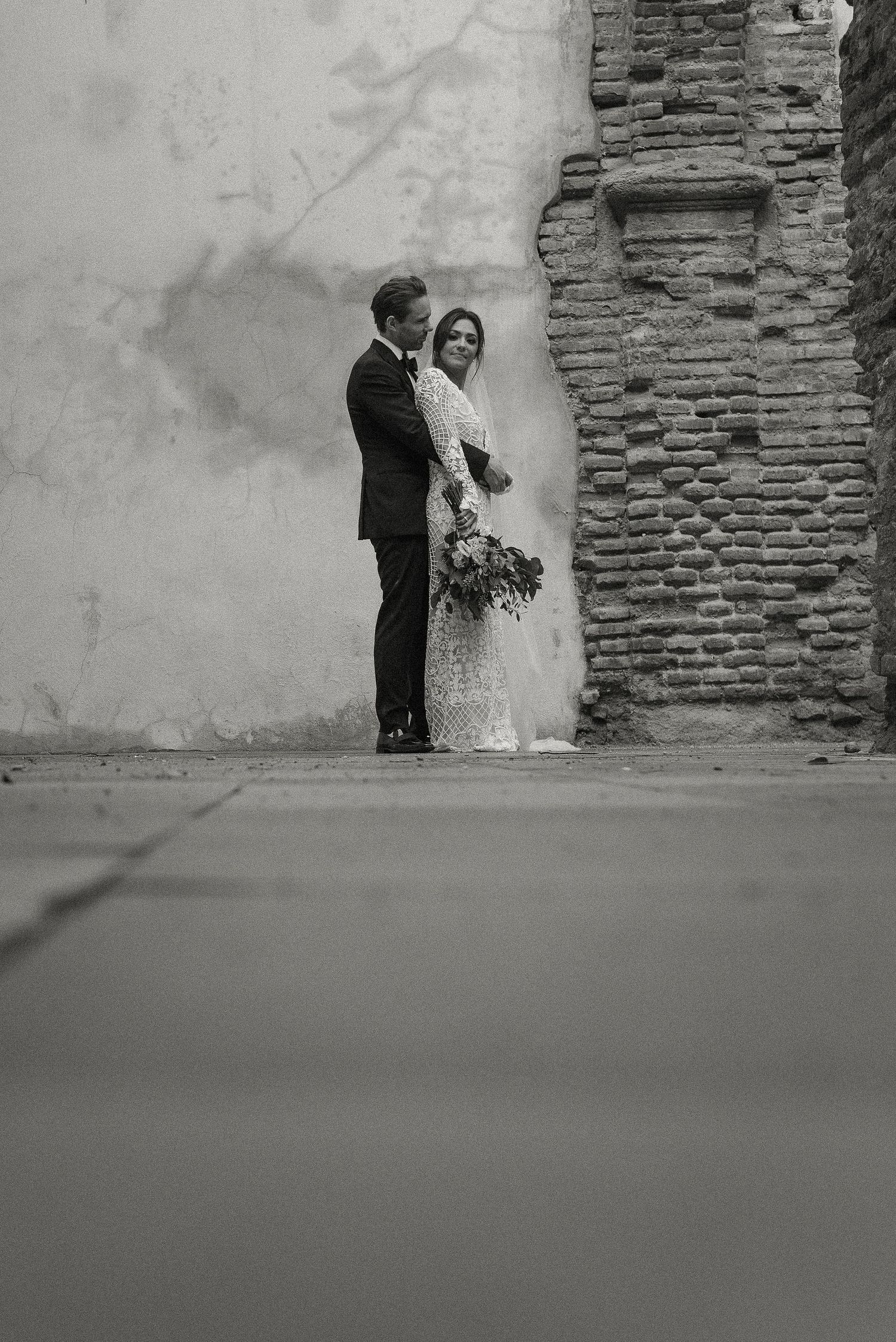 A black and white wedding photo of couple standing against a rustic stone wall with shadows and beautiful bouquet.
