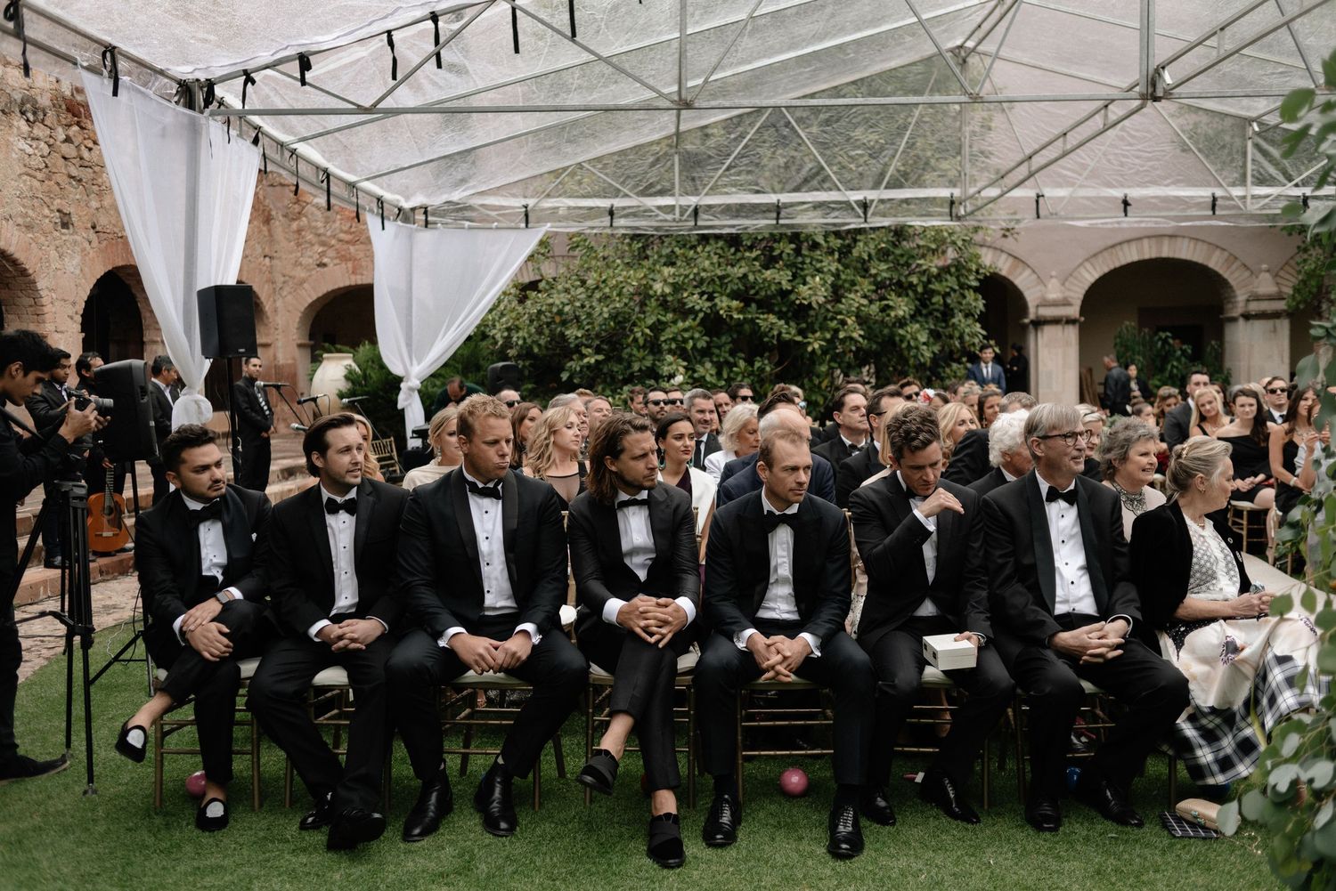Wedding guests in formal black tuxedos sit in rows during an outdoor garden ceremony under a white tent.